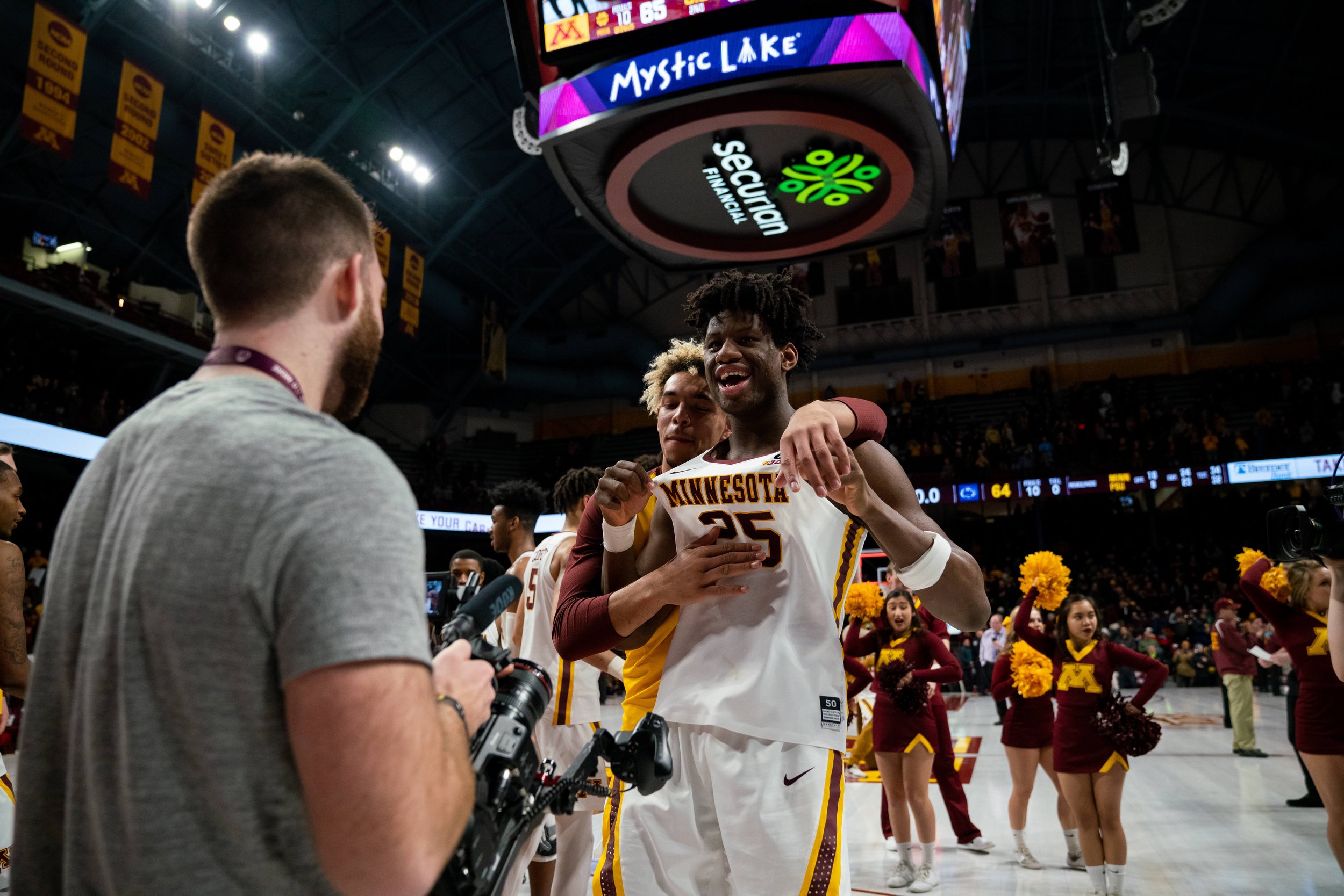 Basketball players celebrating on court with Minnesota jerseys, surrounded by cheerleaders and a photographer, under a scoreboard in an arena.