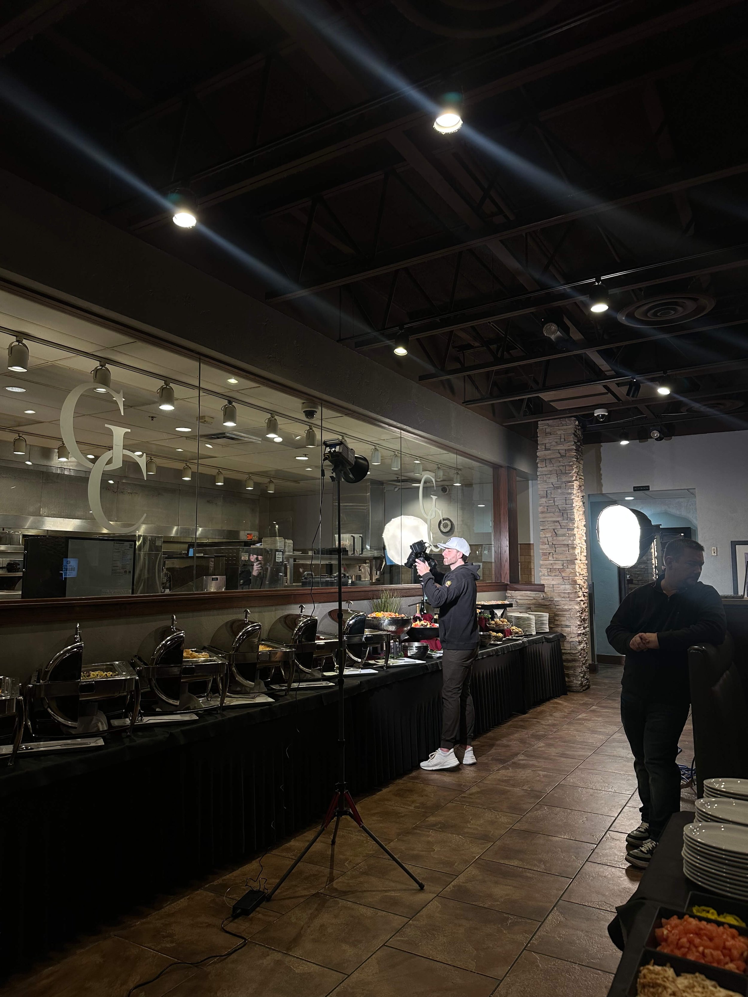 A photographer is taking pictures of a buffet setup in a restaurant. The buffet includes chafing dishes and a variety of food items. The restaurant has a modern industrial design with exposed ceiling beams and stone accents. Another person is standin