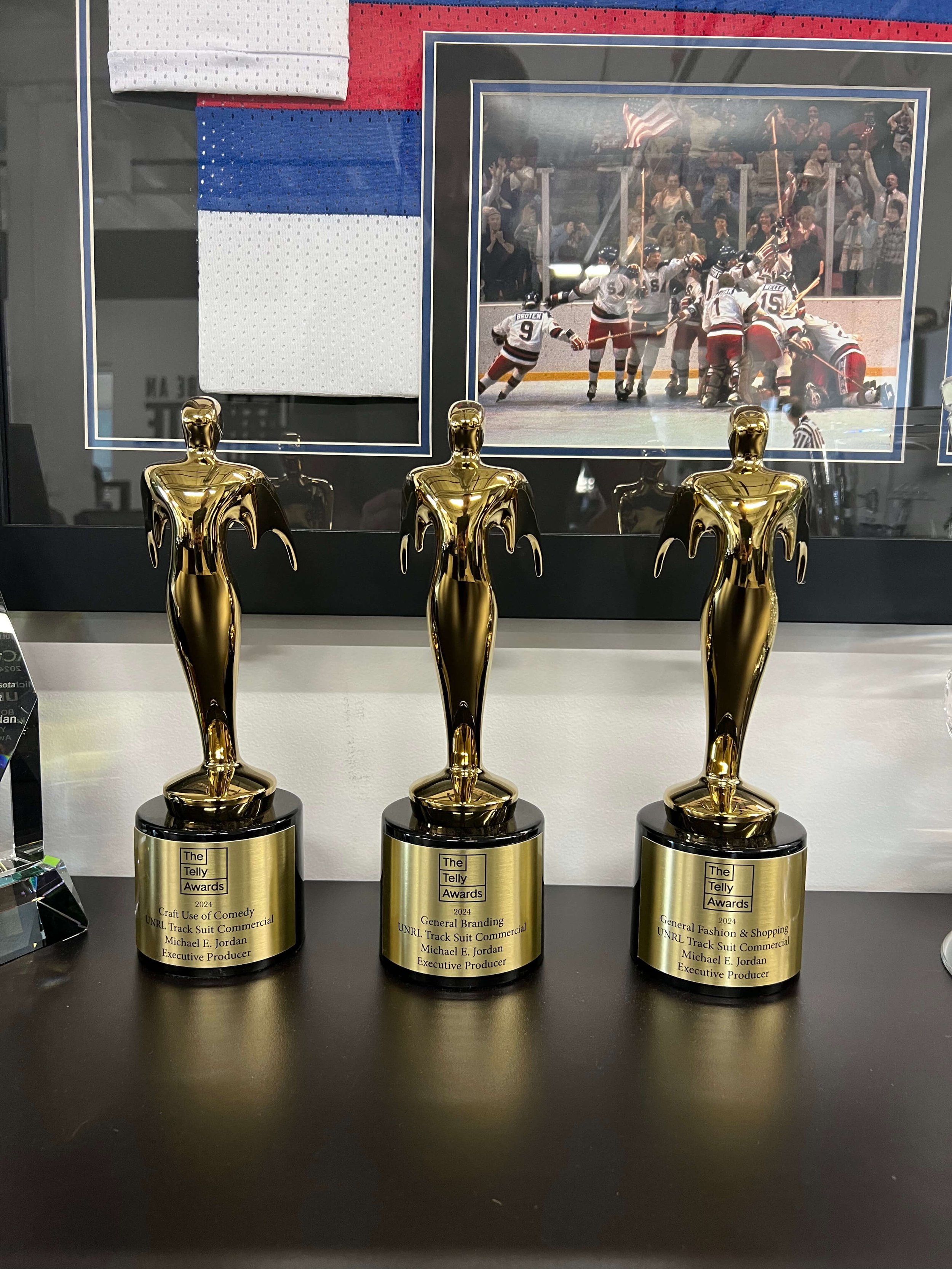 Three Telly Awards trophies on a shelf, with a framed hockey jersey and photograph in the background.
