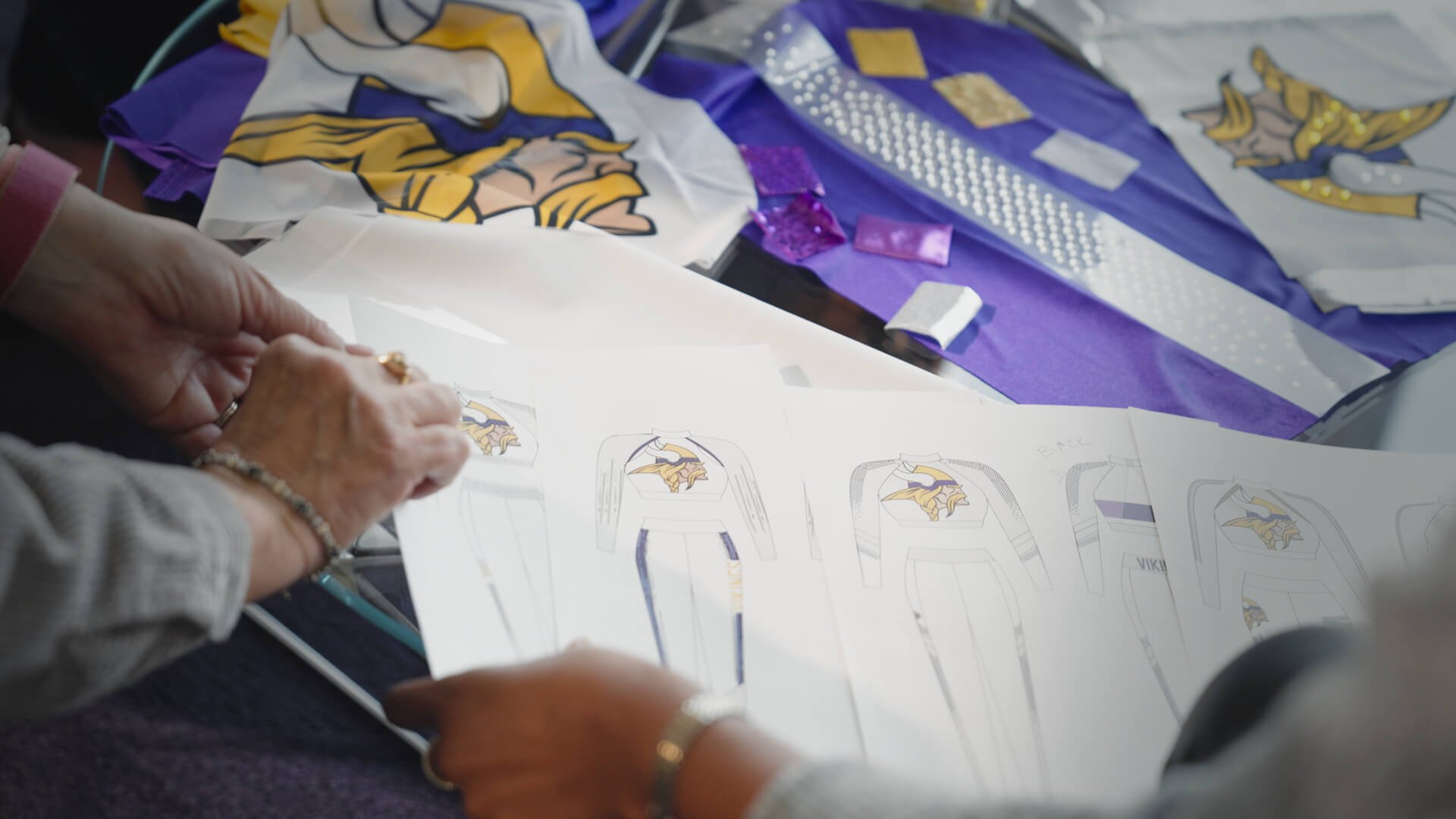 Design sketches of sports uniforms with a Viking logo, surrounded by purple fabric and embellishments.
