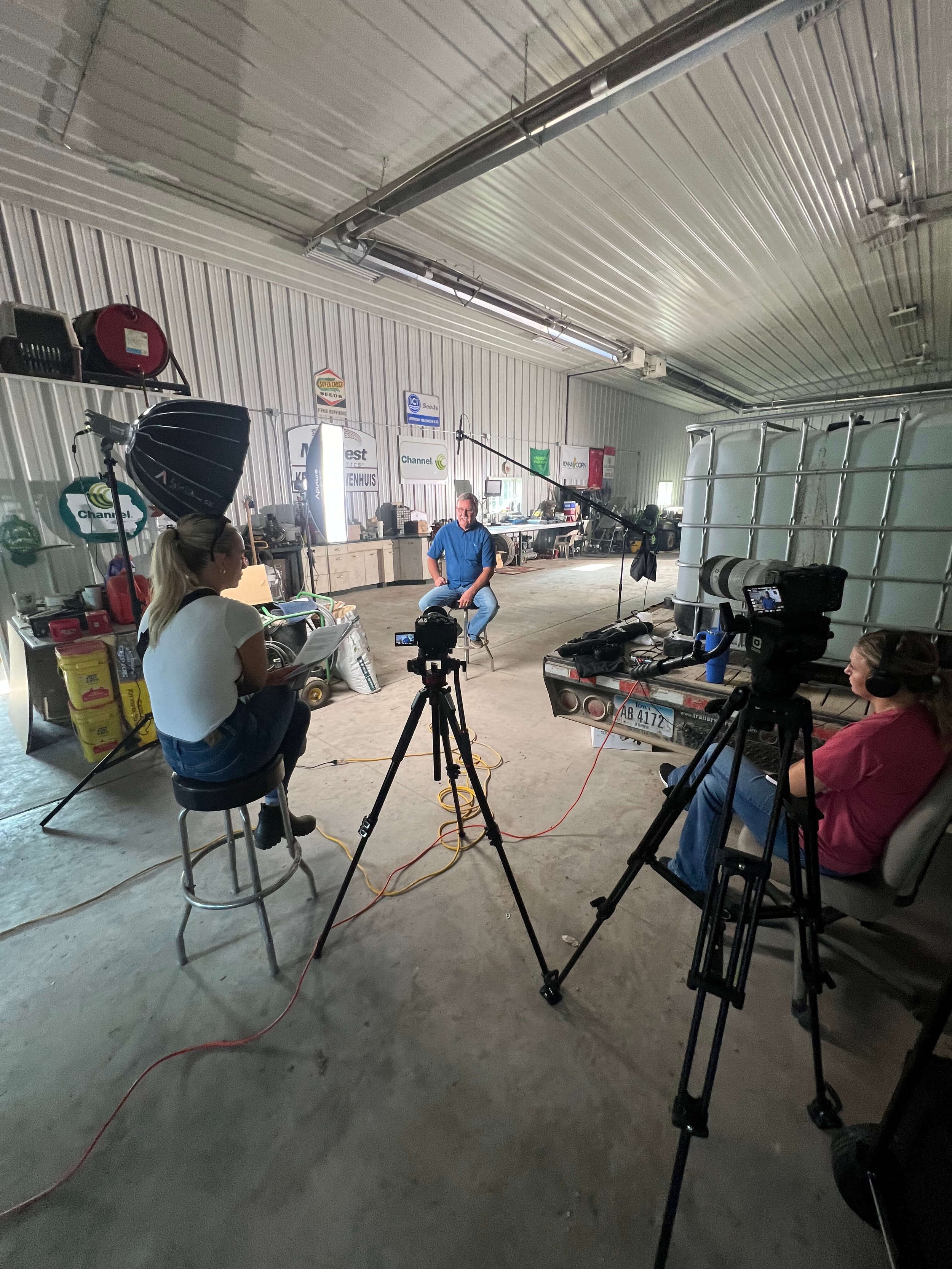 Indoor interview setup in a garage with two cameras, lighting equipment, and three people, one being interviewed.