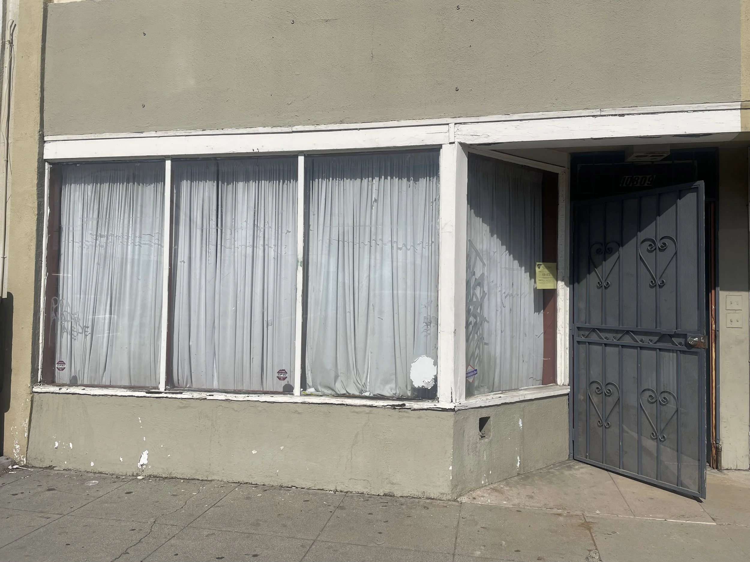 A storefront with large windows covered by white curtains and a black wrought iron gate entrance, with a small yellow sign on the wall next to the gate.