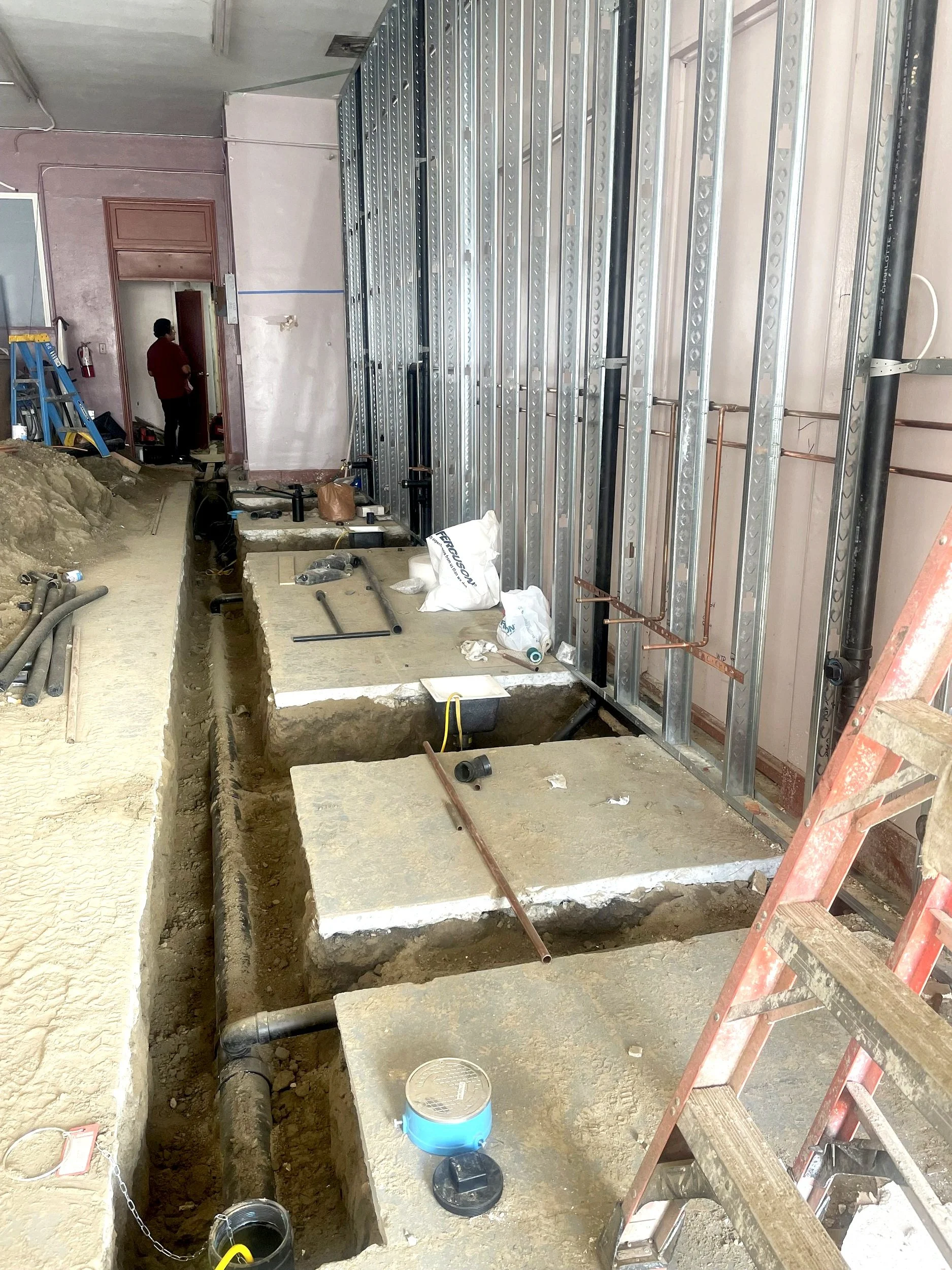 Construction site of interior building with metal wall framing, pipes installed in the floor, and a ladder on the right side. Construction tools and materials are scattered on the floor.