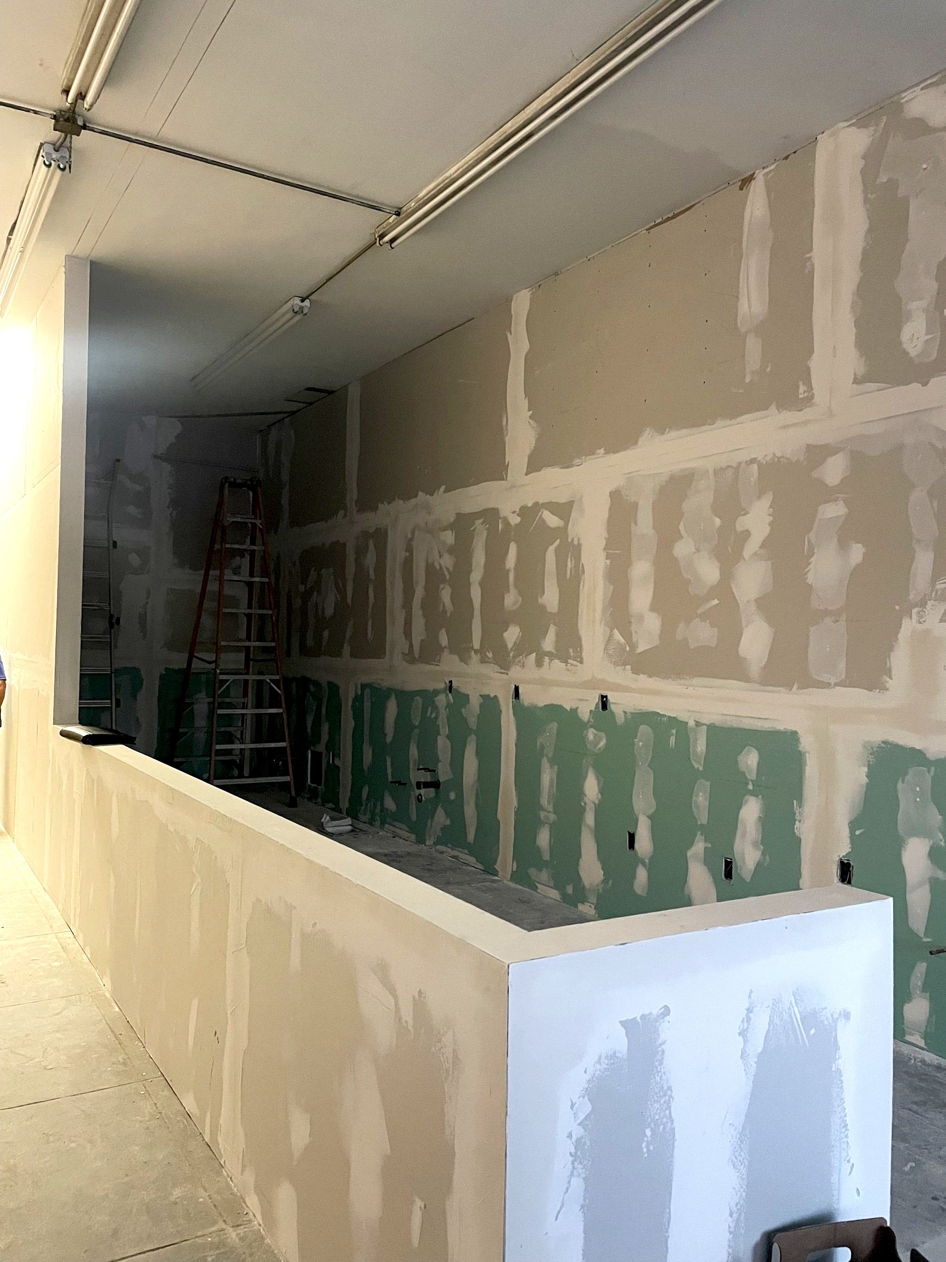Partially constructed interior wall with drywall installation, green drywall on lower part, ladder, and unfinished ceiling with lighting fixtures.