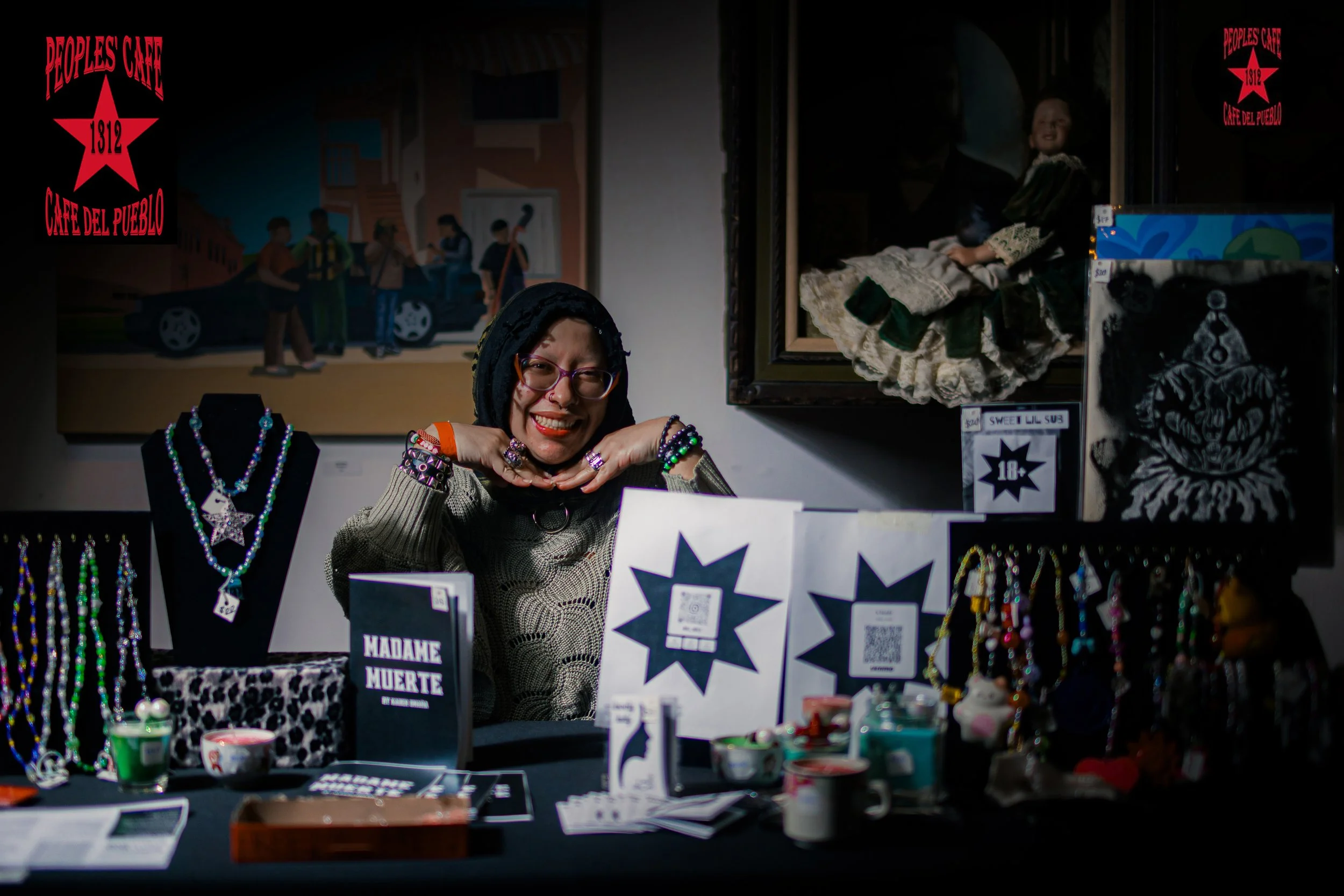 A woman with glasses and long dark hair, smiling and posing with her hands under her chin at a jewelry booth. The booth displays necklaces, bracelets, and other accessories, with a sign reading "Madame Muerte" and other items like QR codes on the tab