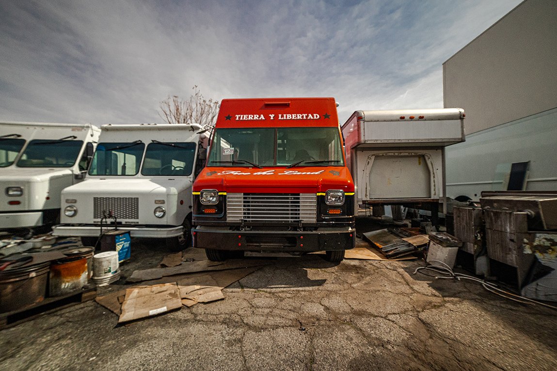 A row of old commercial trucks and vehicles parked outdoors on a cracked asphalt surface, with cloudy sky and a building in the background.