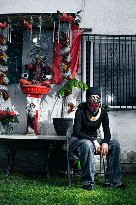 Person sitting on a chair outside next to a decorated altar with religious figures and flowers, wearing dark clothing, a black mask with a red design, and a black beanie.