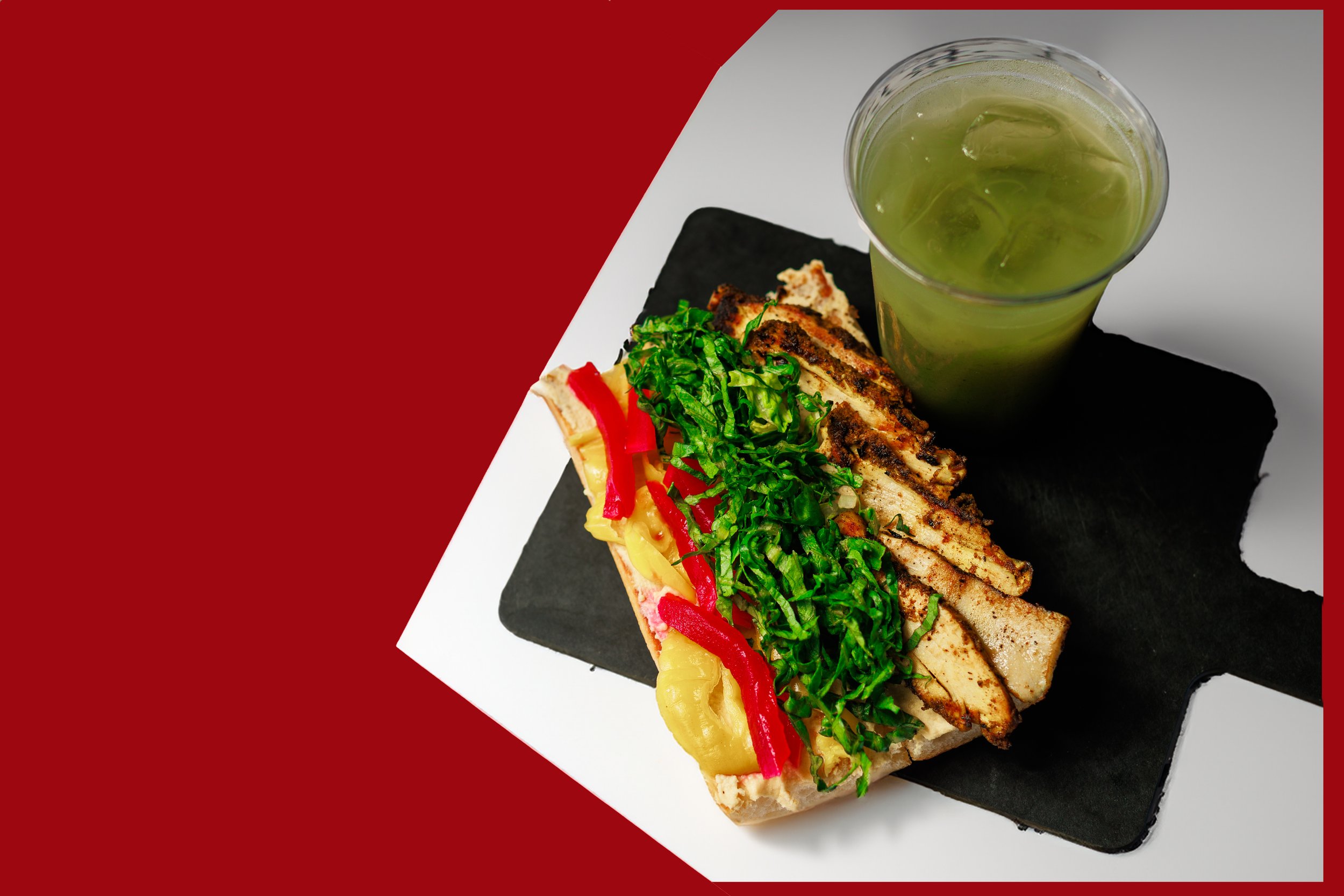 Grilled chicken sandwich with lettuce, red bell peppers, and pineapple on a bun, served with a side of green juice with ice cubes on a black slate and white surface with a red background.