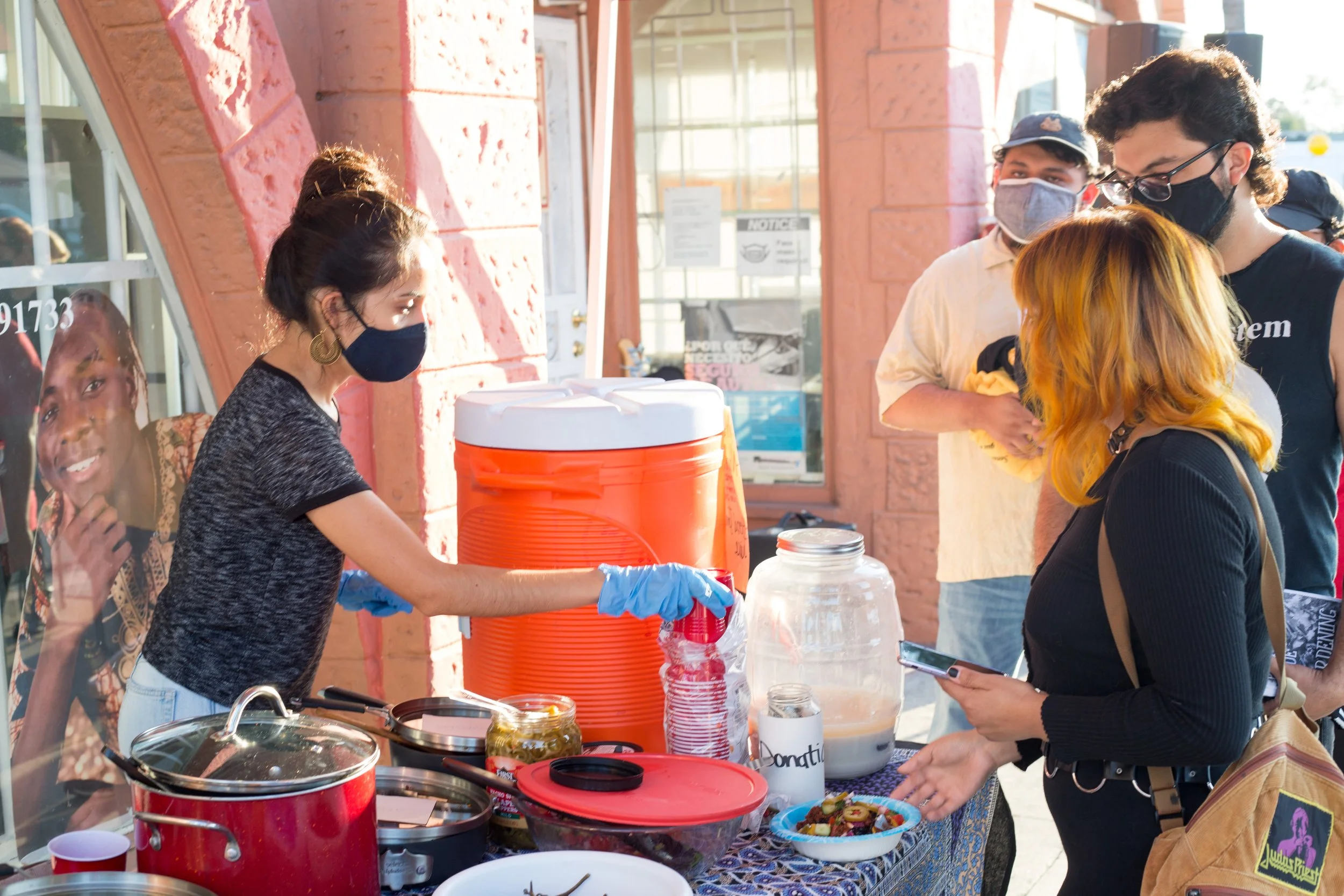 A group of people at an outdoor food stand, with a woman serving food wearing a black face mask and blue gloves, while a woman with orange hair and a black shirt is holding a phone and a bowl of food, and two men in the background wait in line, all w
