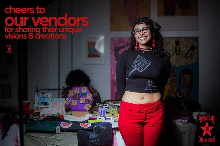 Smiling young woman with curly hair and glasses standing at a vendor booth with various handmade crafts and artwork on display, during an event supporting vendors and sharing creative visions.