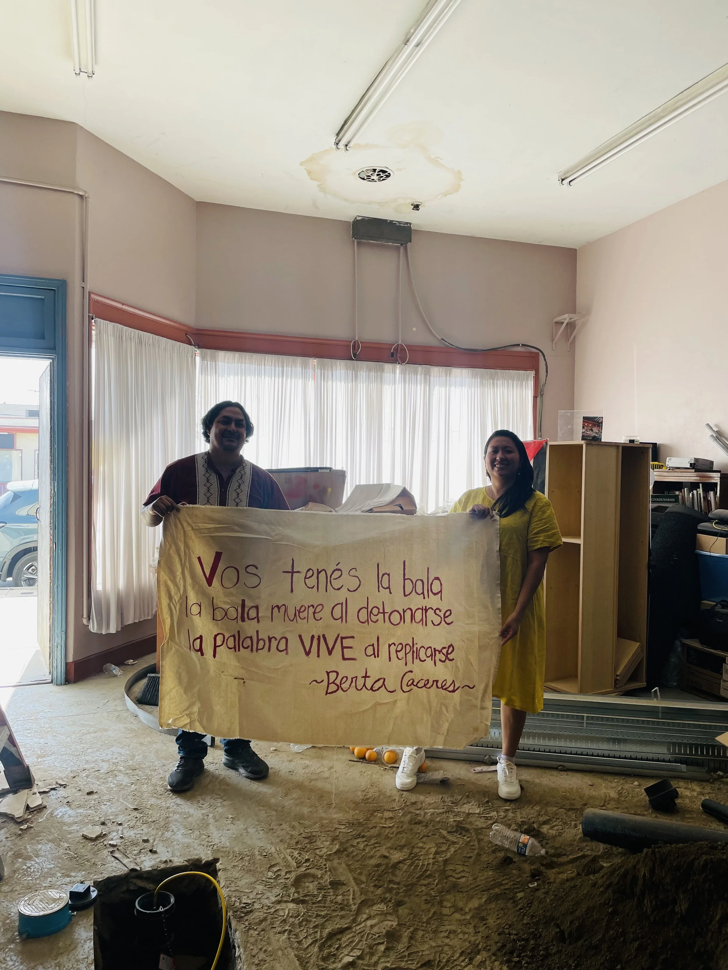 Two people holding a banner inside a room with construction debris on the floor, a pile of books and appliances on the shelves, and sunlight coming through sheer curtains.