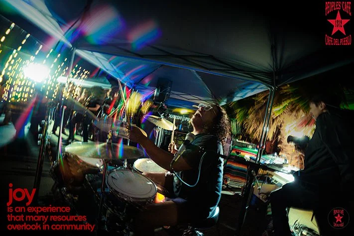 A drummer and a keyboardist perform at an outdoor event or party during the night, with colorful lights and a canopy overhead. In the background, there is a screen displaying lights, and a quote in red text overlays the scene.