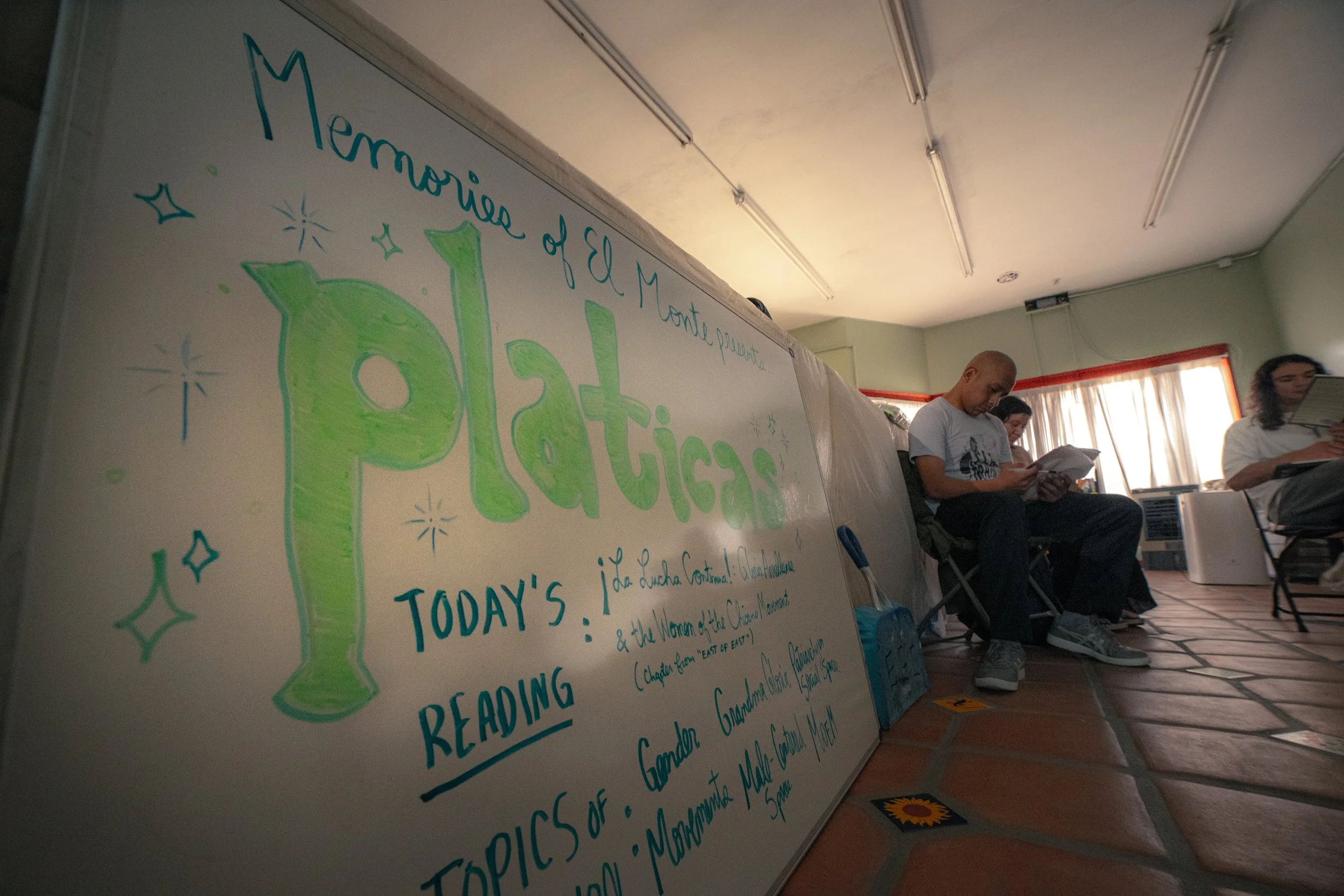 A whiteboard with handwritten text about El Mantel de Pláticas, displaying today's reading topics, with several people sitting on chairs reading papers in a room with a tiled floor and curtains on the window.