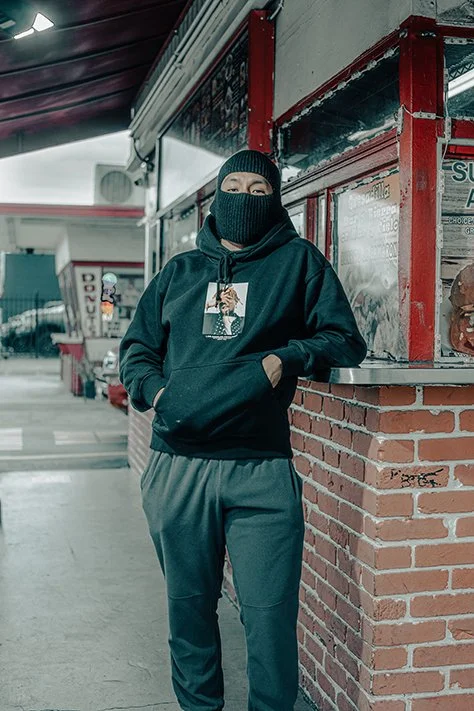 Person wearing a black hoodie with a graphic print, black face mask, and dark sweatpants, standing outside a storefront.