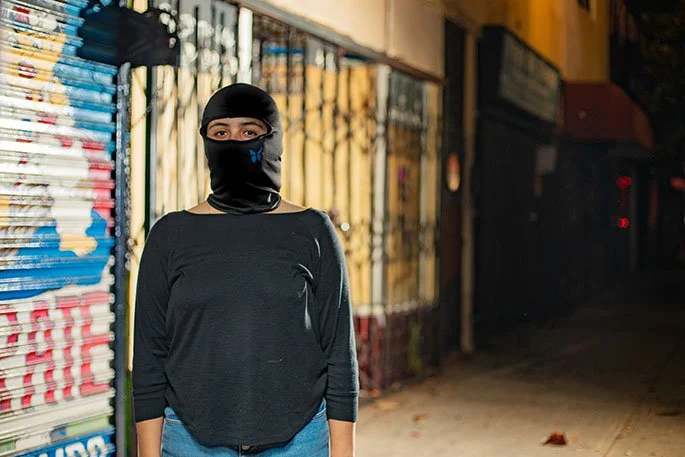 A woman wearing a black face mask and black long-sleeve shirt standing on a sidewalk at night, with a metal fence and store windows behind her.