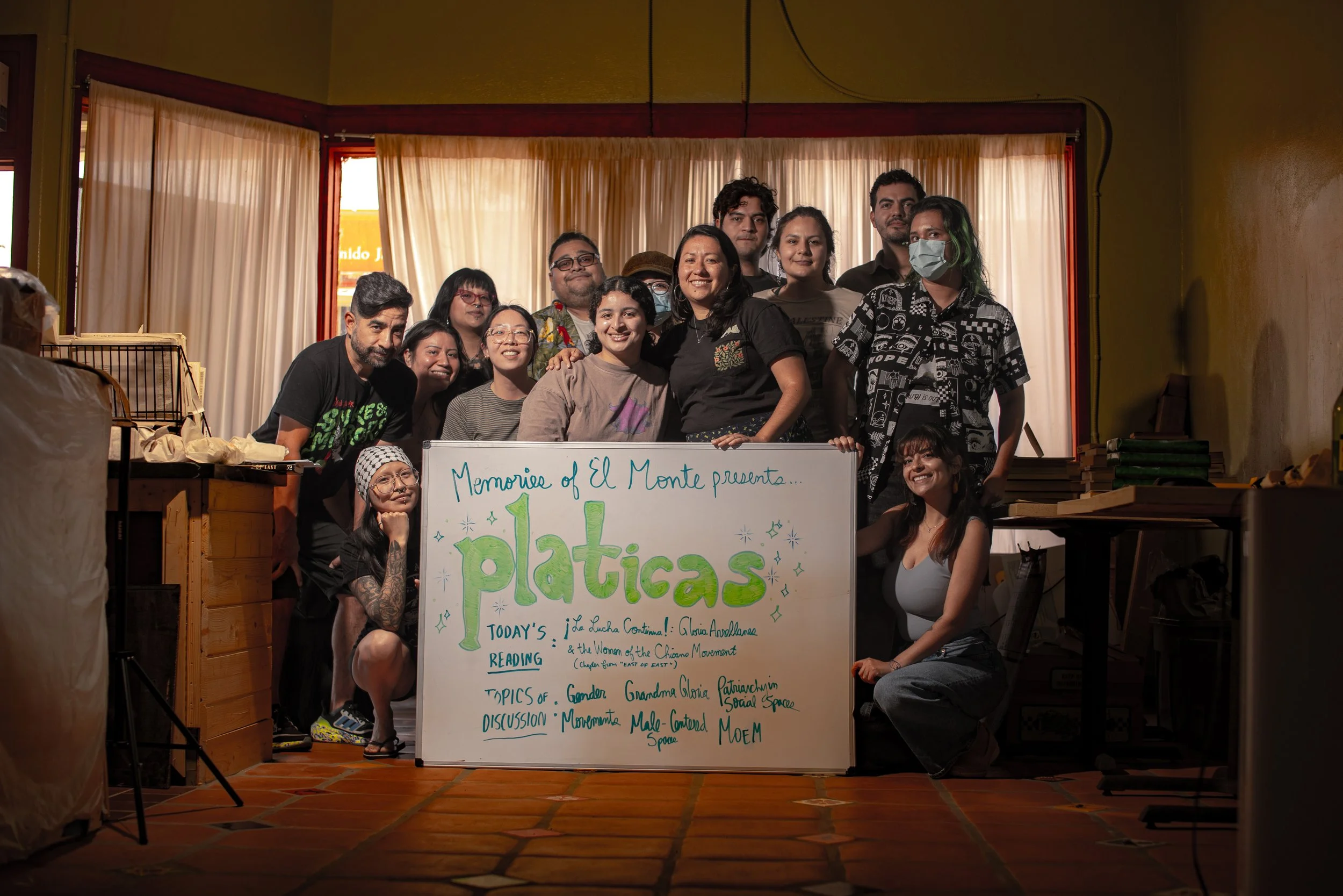 A group of people gathered around a whiteboard with colorful writing, standing indoors in front of a window with beige curtains. The group appears to be at an event or workshop, smiling at the camera.