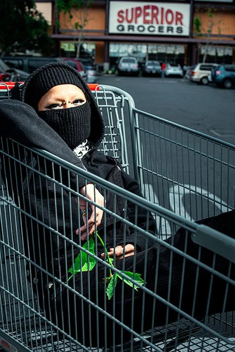 A person wearing black clothing, a face mask, and a beanie sitting in a shopping cart outside a grocery store named Superior Grocers, in a parking lot with cars.