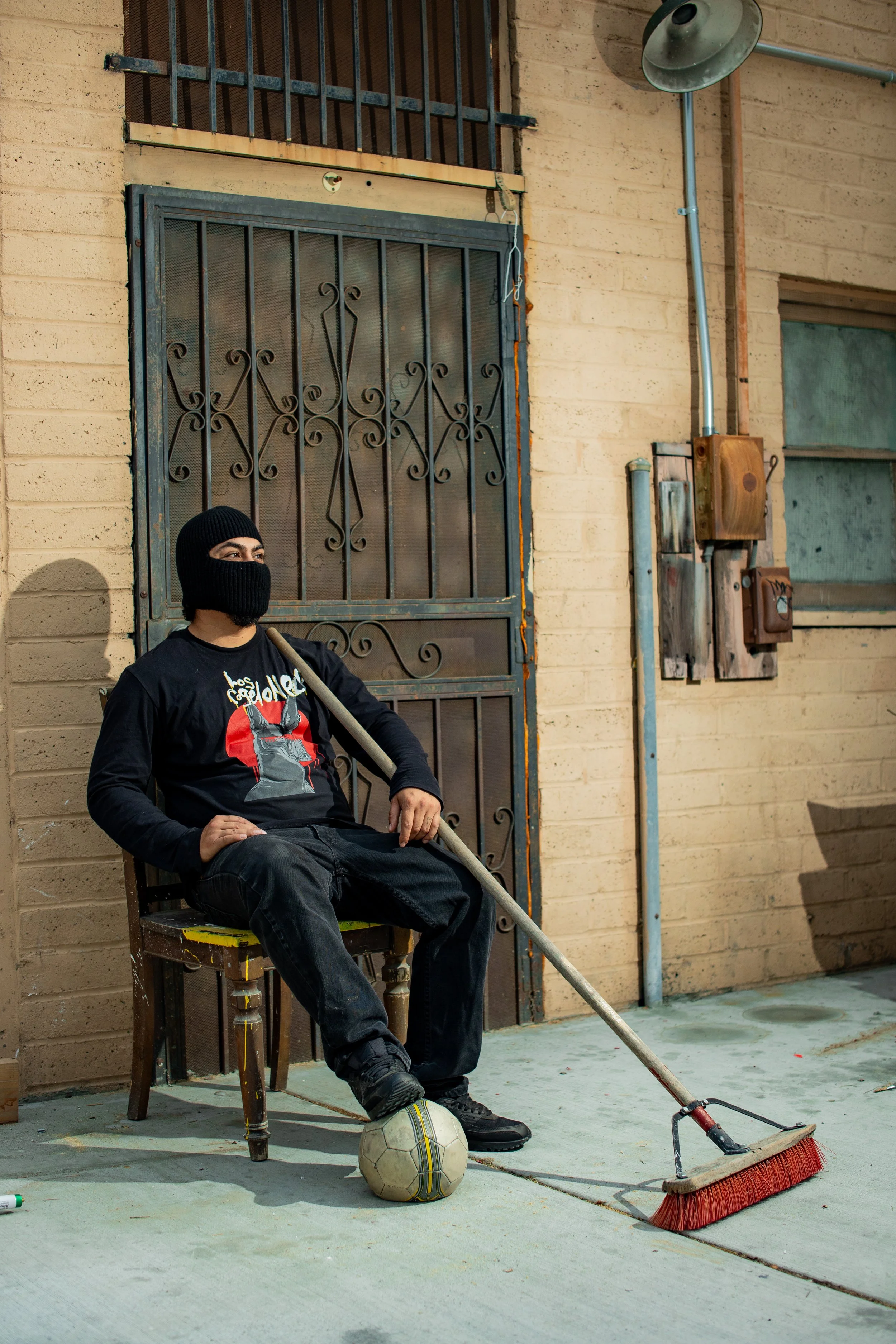 A man wearing a black ski mask, black t-shirt, and black pants is sitting on a chair with one foot on a soccer ball. He holds a broom with a long handle and rounded brush at one end. The setting appears to be an outdoor area with a brick wall, a barred door, and electrical boxes.