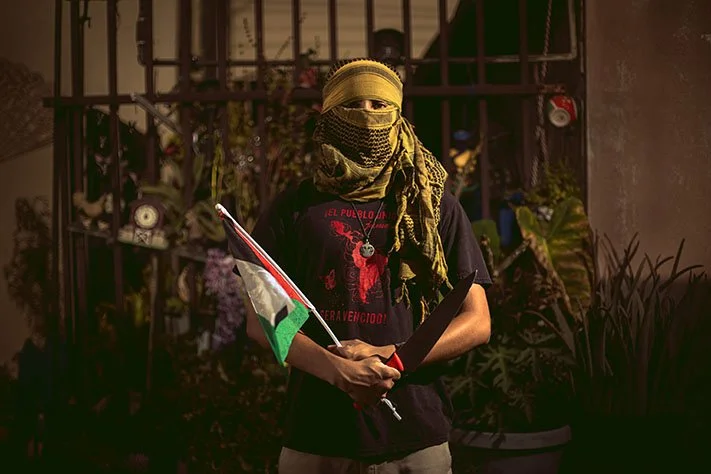 Person with a head covering and face mask holding a flag and a cardboard sign in an outdoor setting with plants and a metal fence.