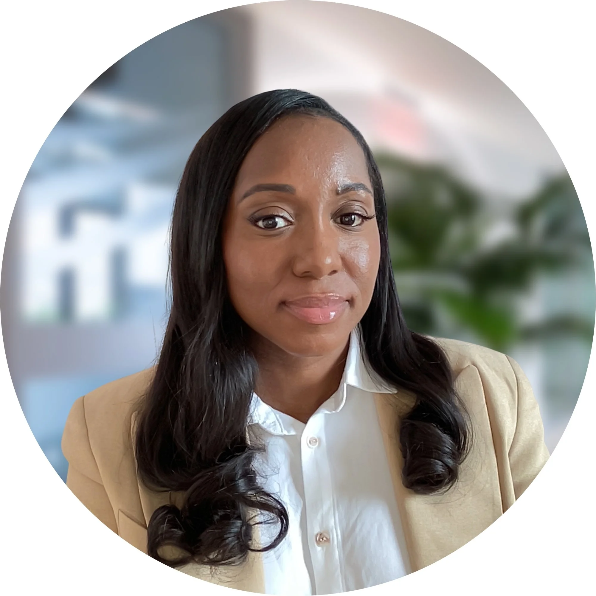 Portrait of a woman with long wavy black hair, wearing a beige blazer and white blouse, in an office setting with blurred background.