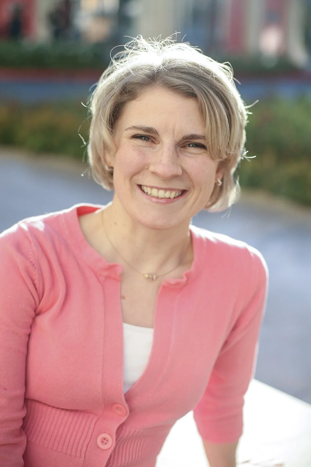 headshots for a broadway actress, the woman is sitting with the sun behind her, she is smiling at the camera wearing a pink sweater and white shirt underneath with short blonde hair