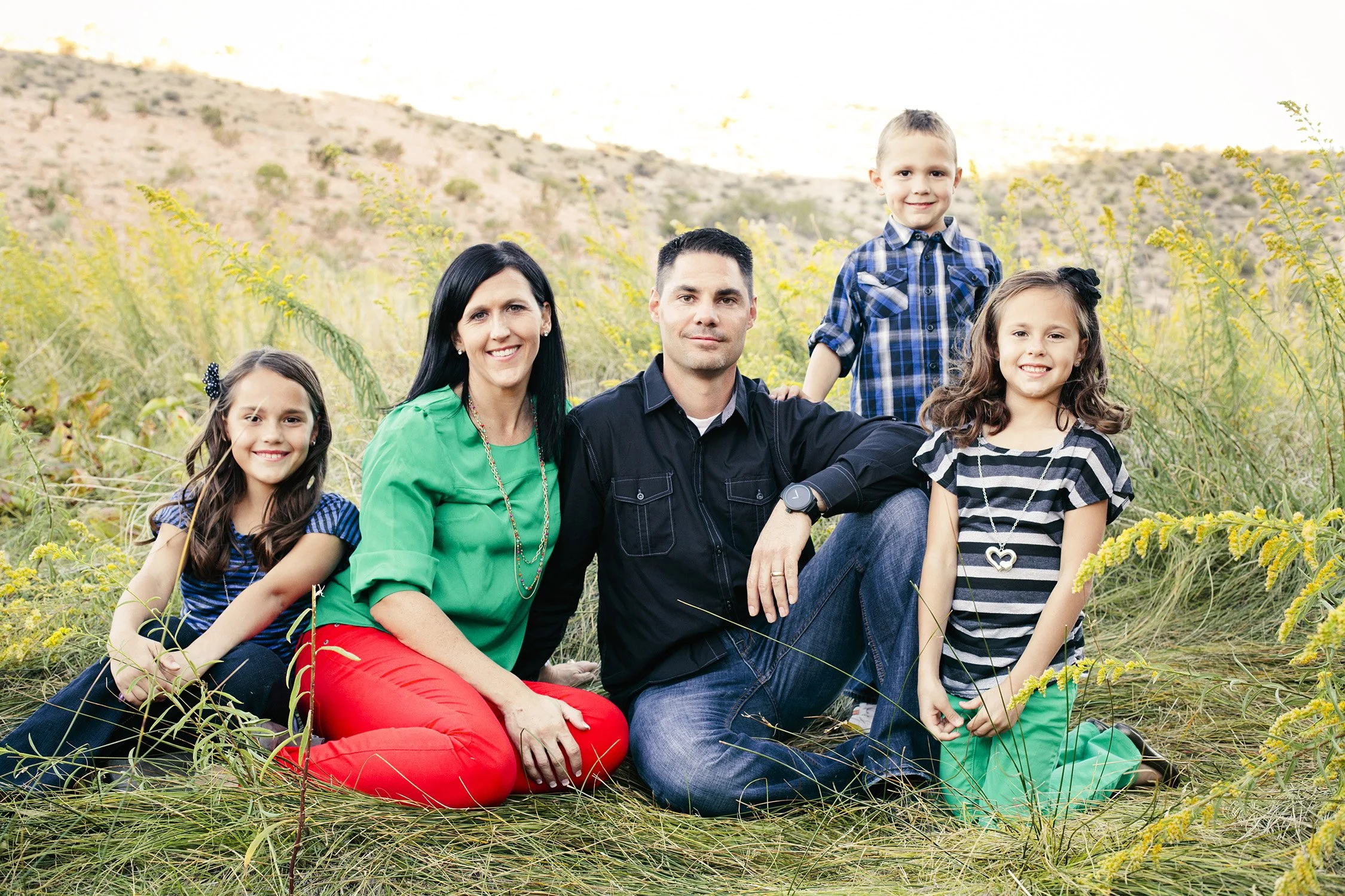 A family of five is sitting on long grass and high shrubs around them. They are smiling at the camera, with mom and dad in the middle, the youngest son standing next to dad, and the two daughters sitting on either side of dad
