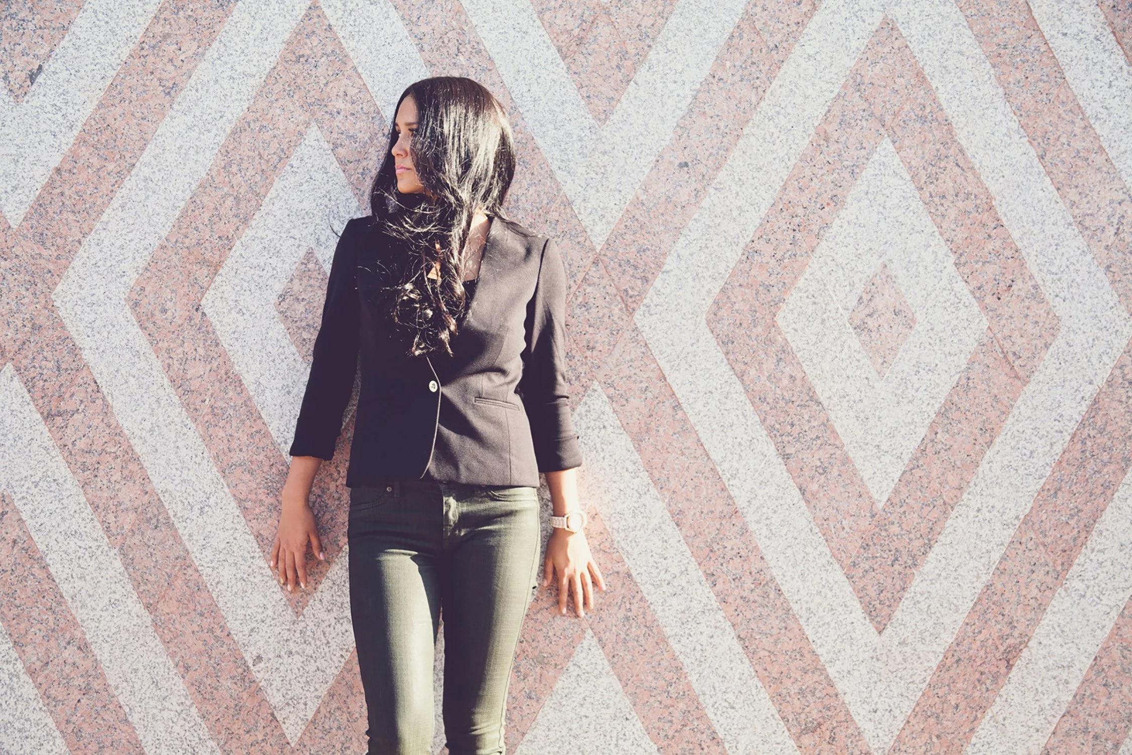 High School senior, Ashley is leaning up against a wall. The wall is white and pink in a large diamond pattern. She is wearing all black and is looking to the left.