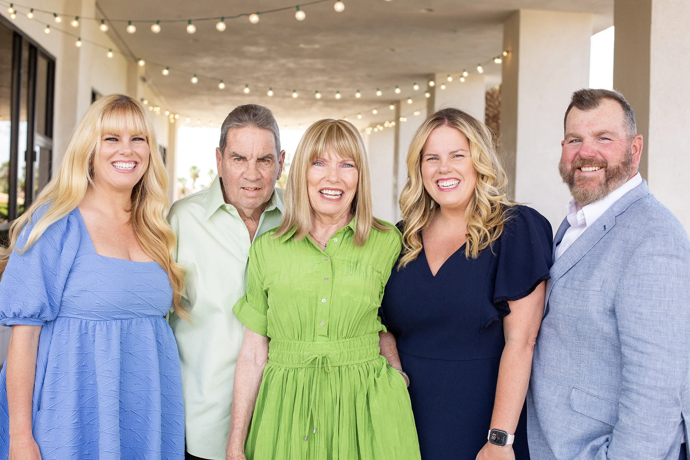 A family of five are standing close together smiling at the camera. The parents are the middle with their grown adult children on each side, the are on a large covered area with string lights above them in the background