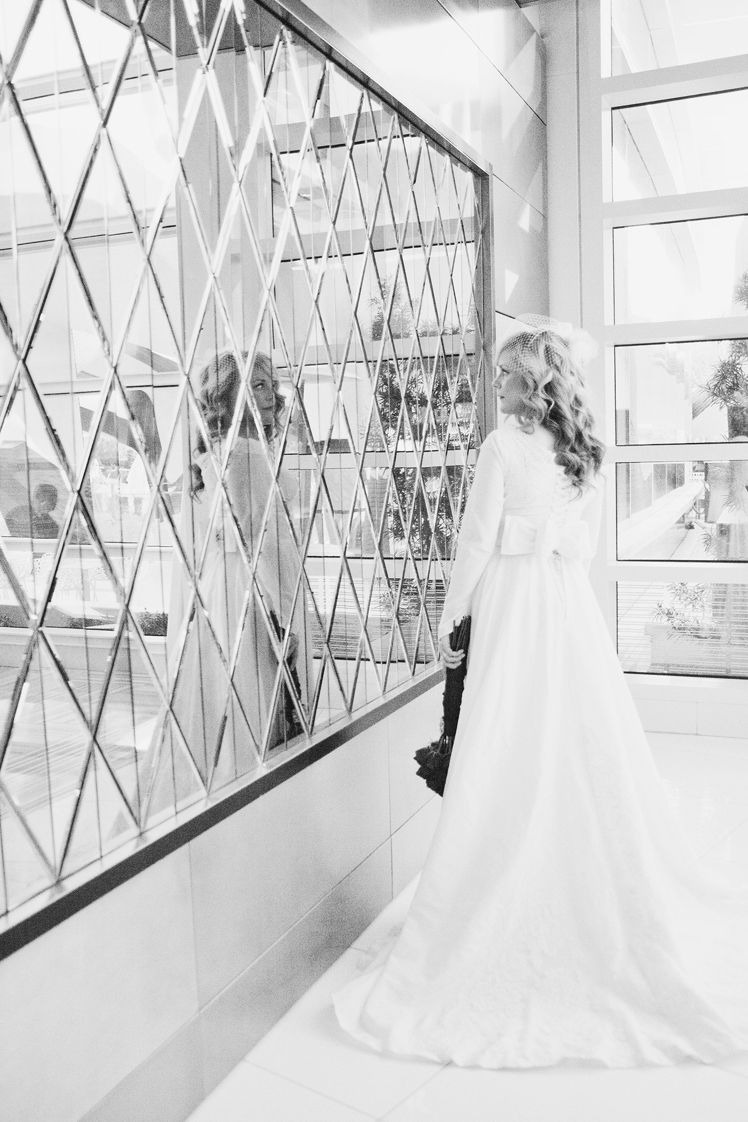 A bride standing in front of a wall of mirrors looking at her reflection, back to the camera to show bridal gown train