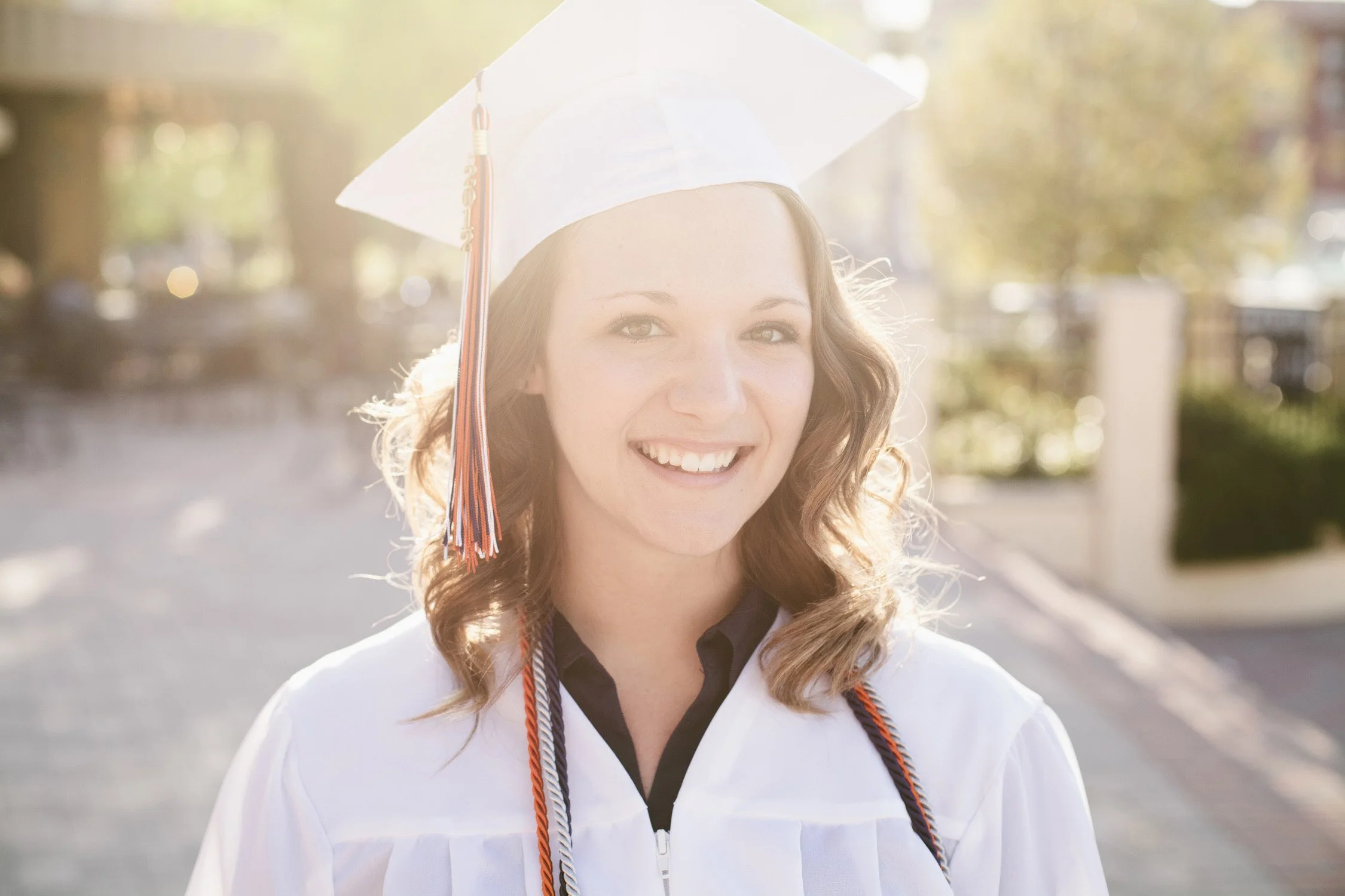 Wearing white graduation wardrobe and hat, a high school senior, is smiling at the camera with the sun shining behind her.