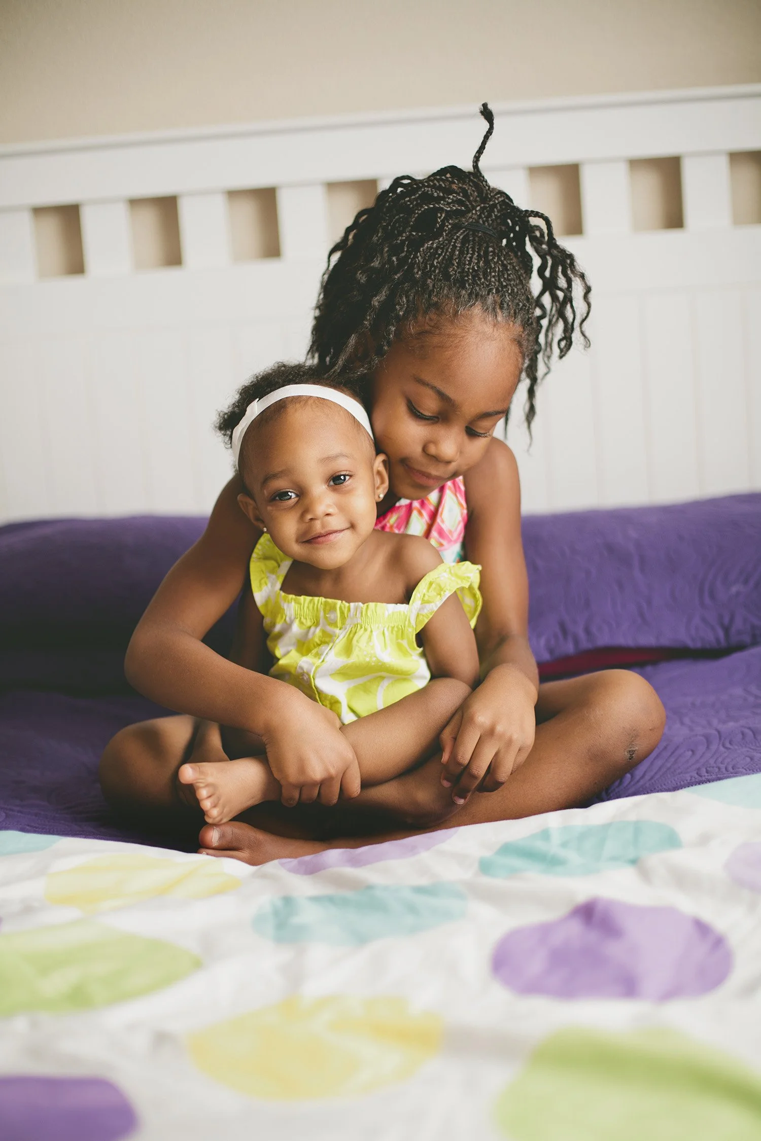 Two young sisters sitting on the bed, the older sister has her baby sister sitting in her lap
