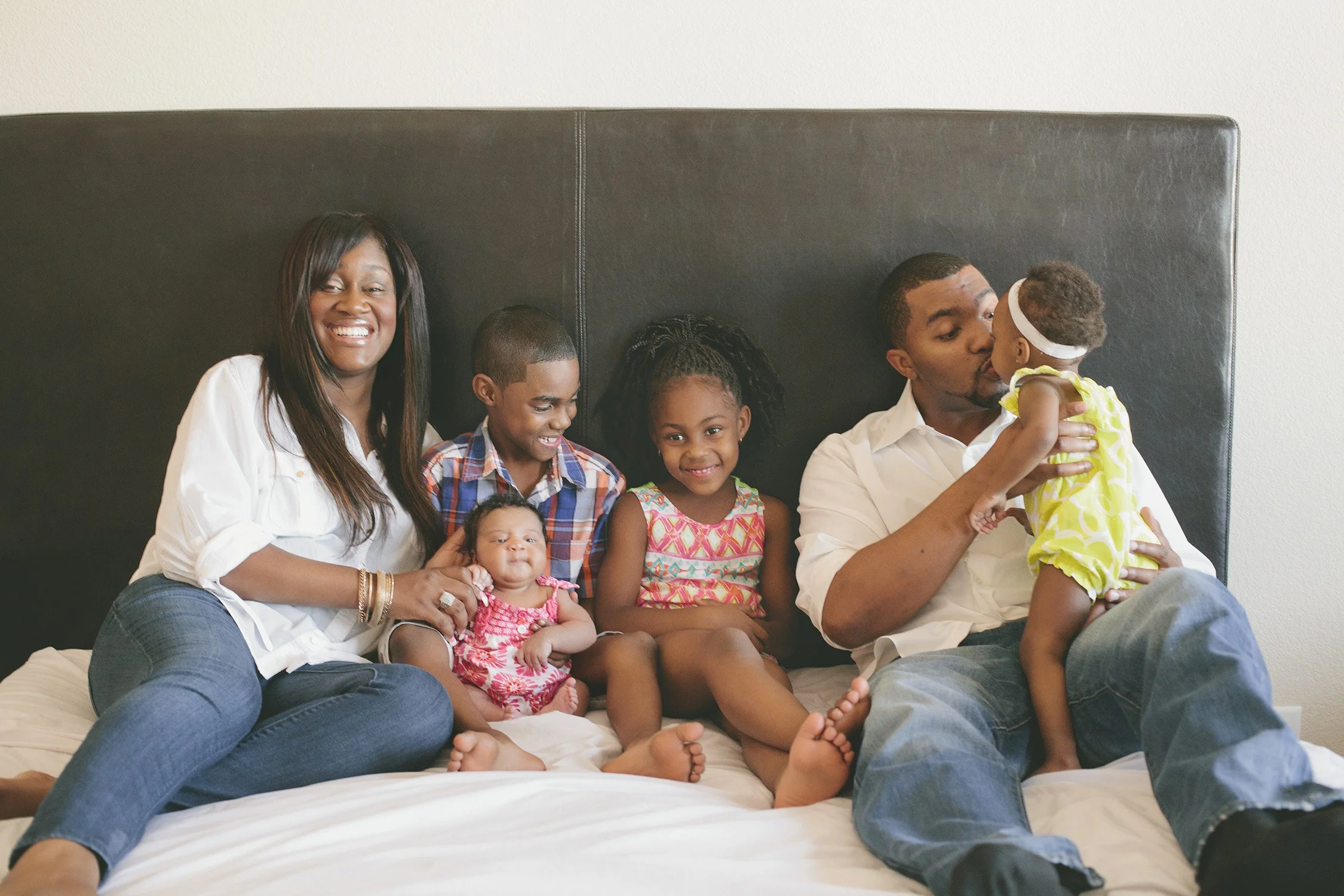 A family of six sitting on a large bed, sitting up against the headboard smiling at the camera, except dad is holding a toddler and giving her a kiss