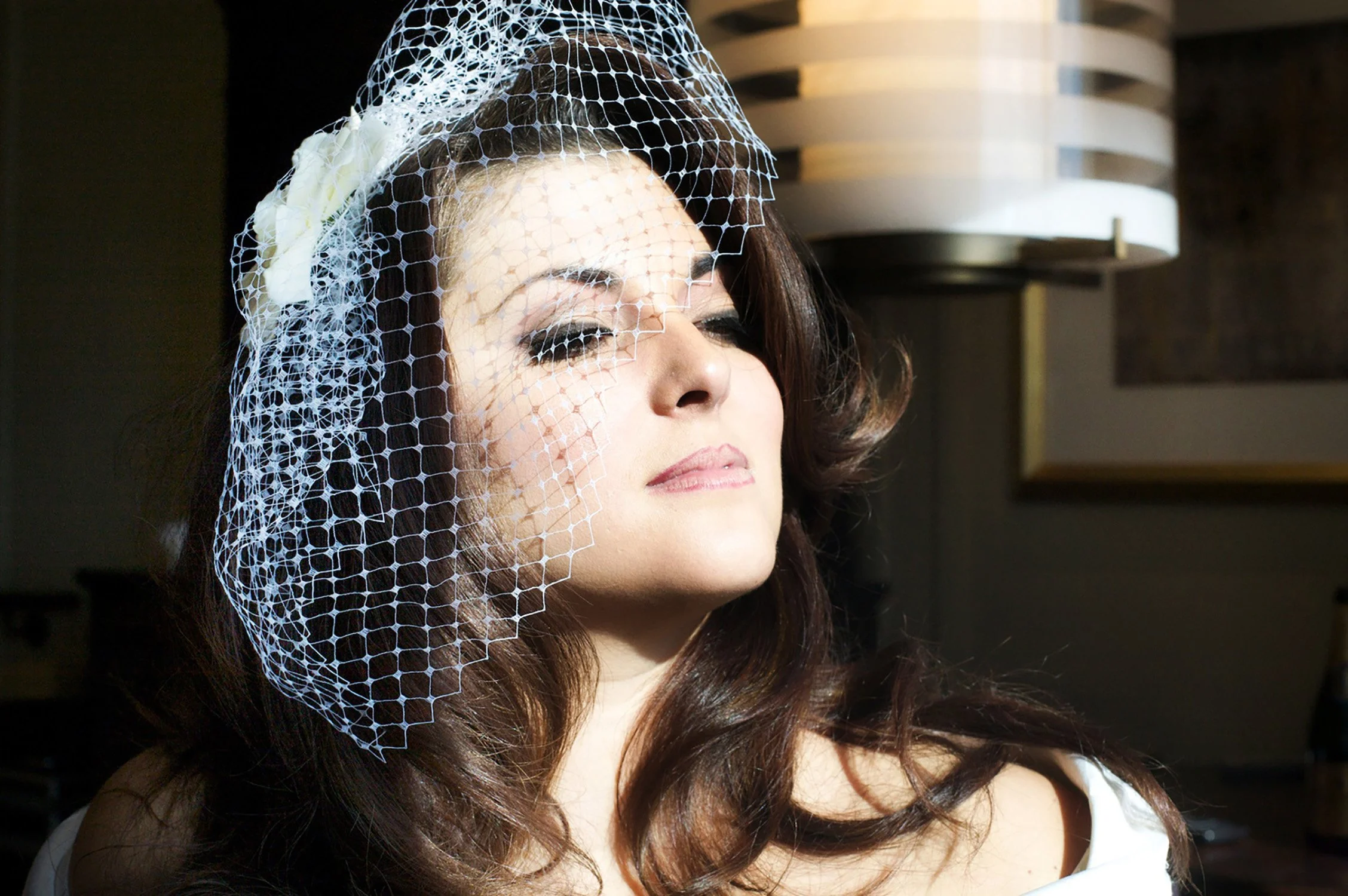 An up close shot of a bride wearing her short net veil, with her eyes closed facing a window before her lipstick is applied