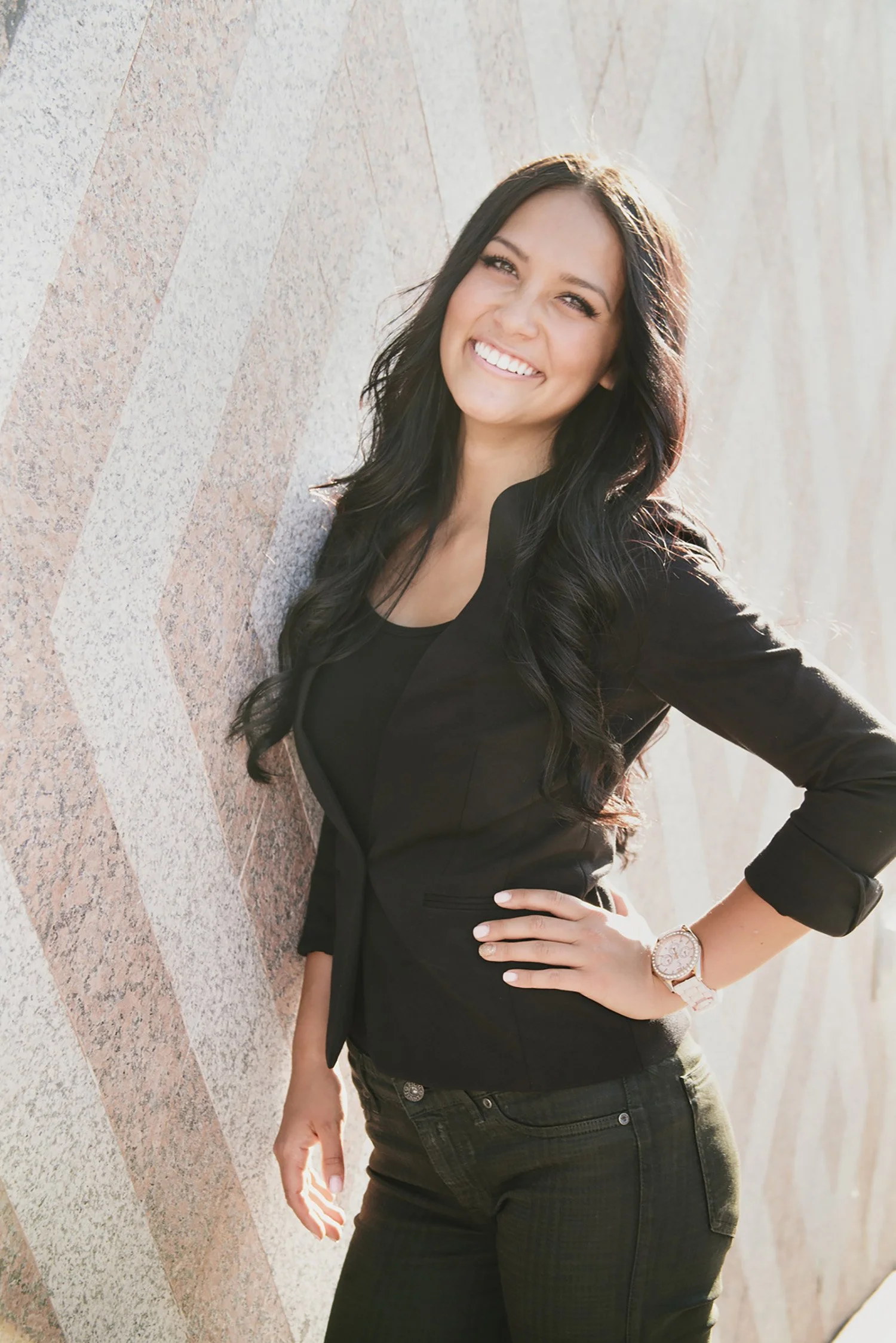 High School Senior, Ashley is leaning one shoulder against a wall with a pink and white diamond pattern made of stone. She is wearing all black. She has one hand on her hip and she is smiling showing a bright smile of teeth at the camera.