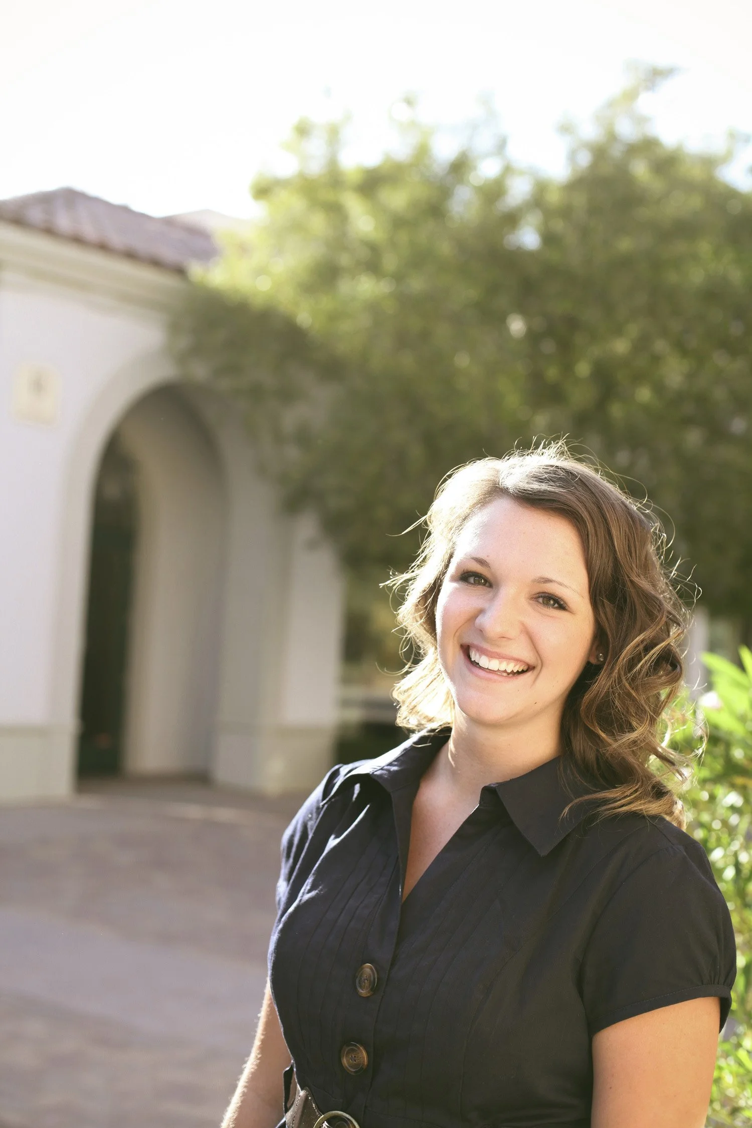 a close up image, the high school senior is smiling at the camera. The background has trees and a rustic building in the background