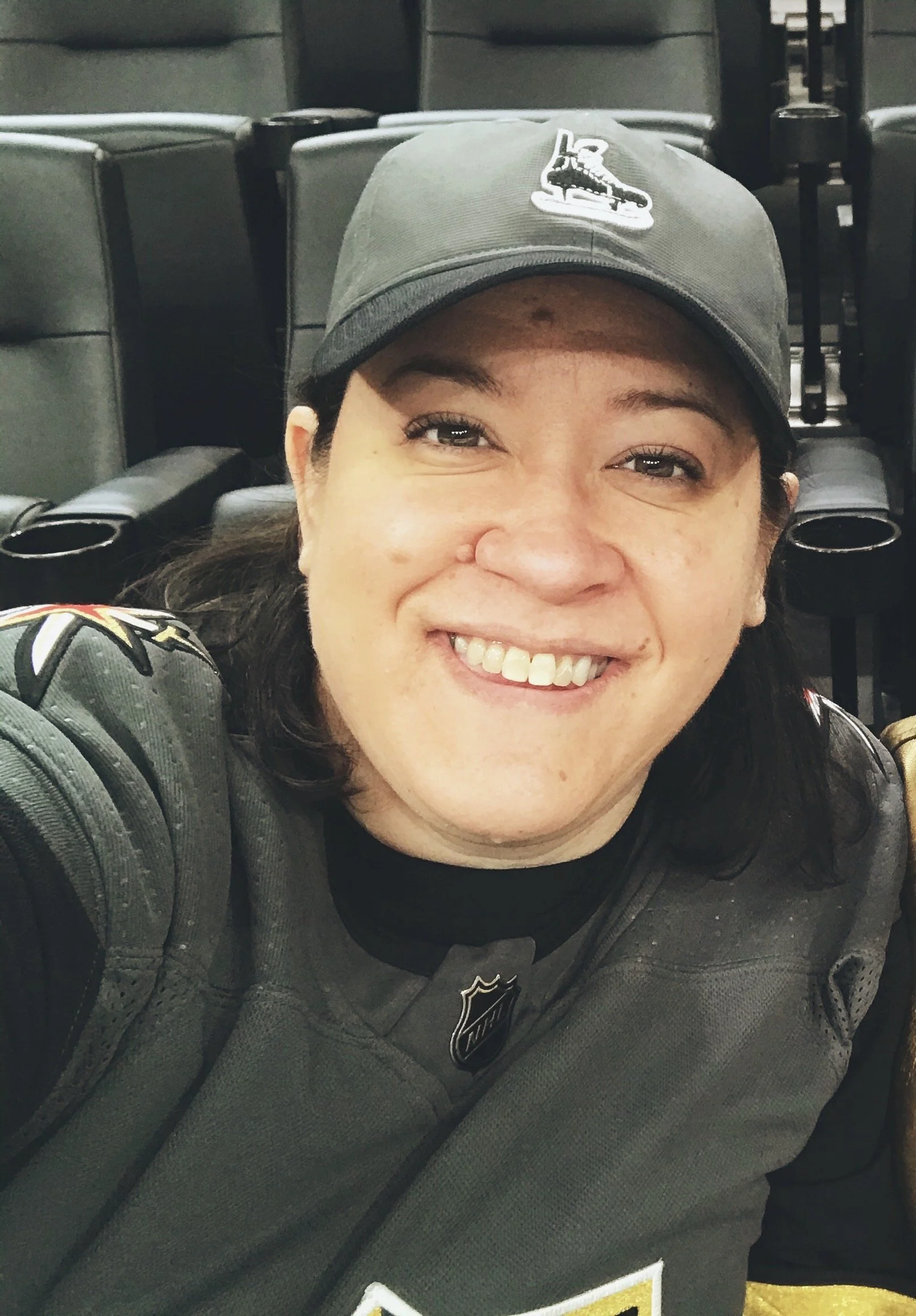 A close up of photographer, Lori Wong. She is wearing a baseball cap with a hockey skate on it, and a hockey jersey with stadium seats in the background. She is smiling at the camera