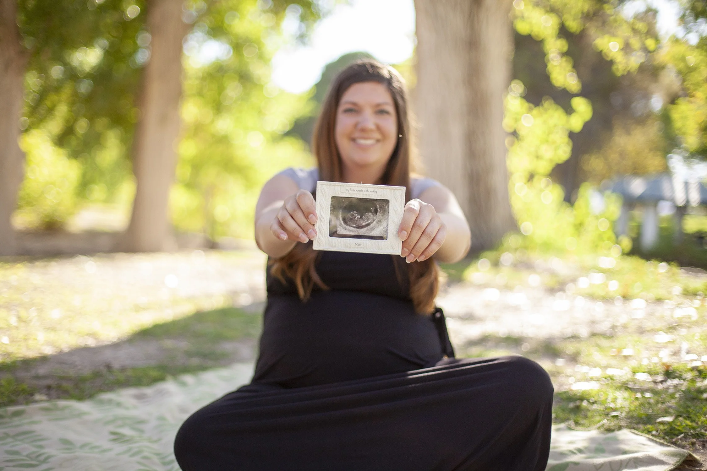 A pregnant woman is sitting on a blanket on the ground, she is holding an ultrasound photo with her arms extended so it's what is in focus the most. There are trees and grass in the background.
