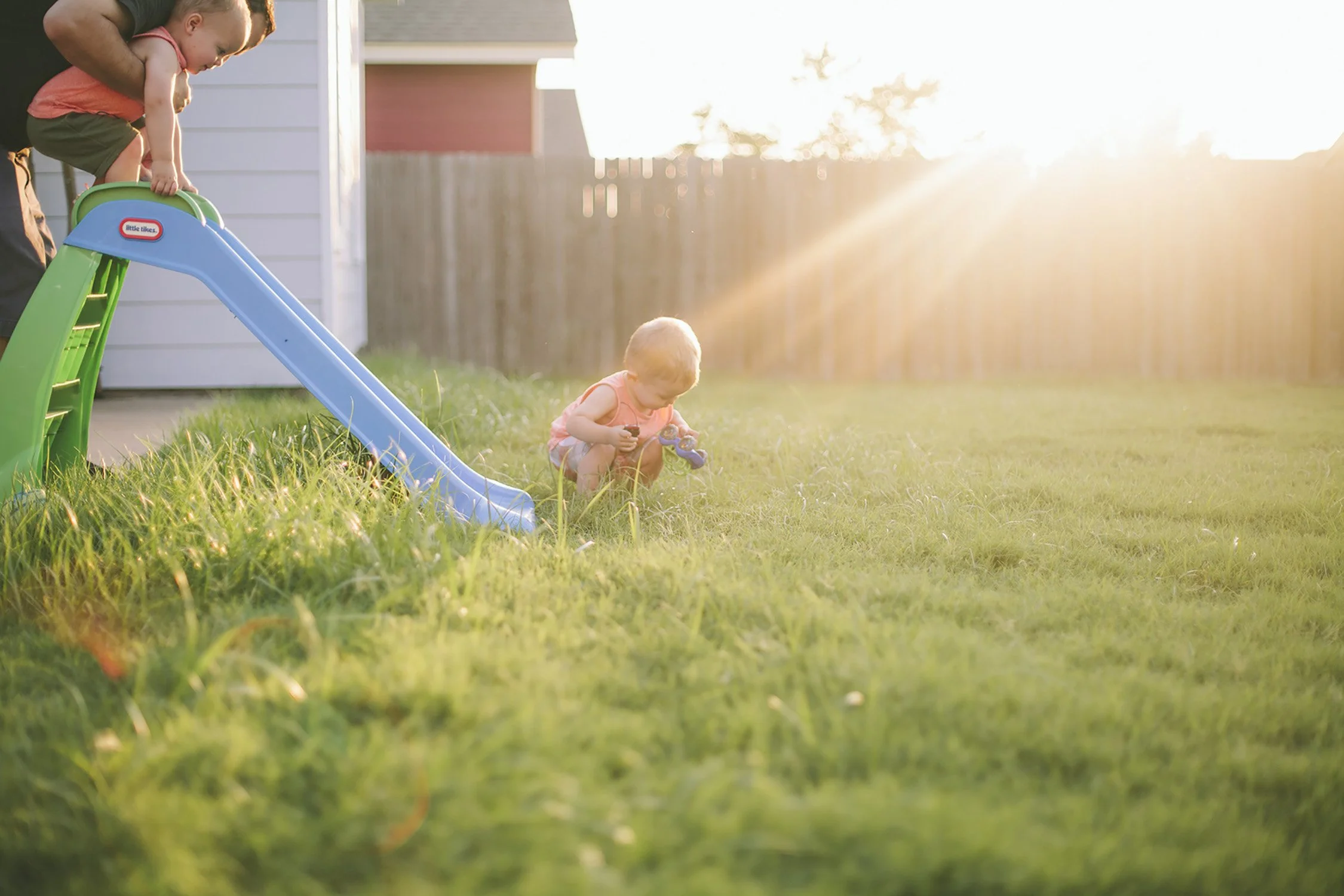 a dad is helping his two sons down a small kid slide in the backyard on the grass. The sun is shining into the camera and onto the family.