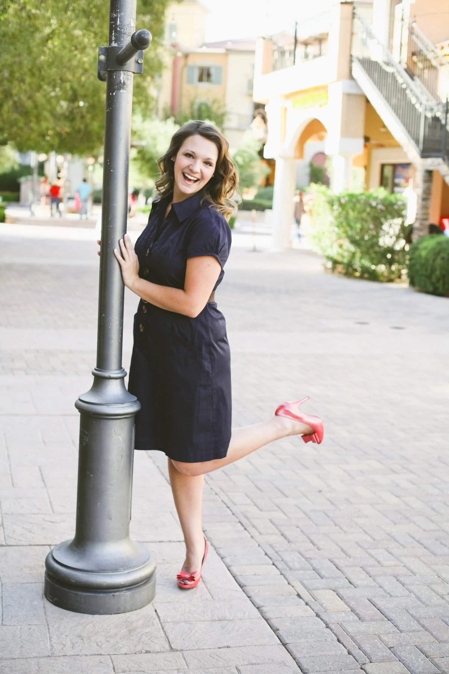 Holding on to a light pole, this high school senior is wearing a navy blue dress and a red high heels, there is an Italy inspired village in the background