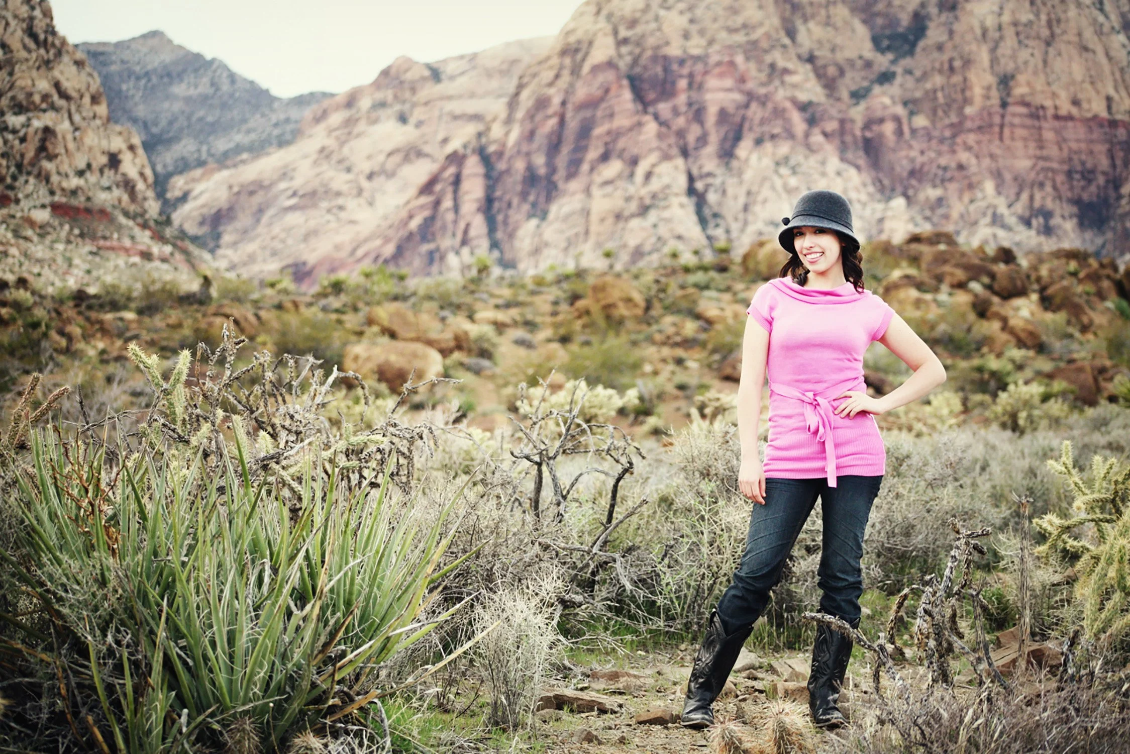 Standing in the middle of the desert and the Red Rock mountains the background, a high school senior smiling and looking at the camera.