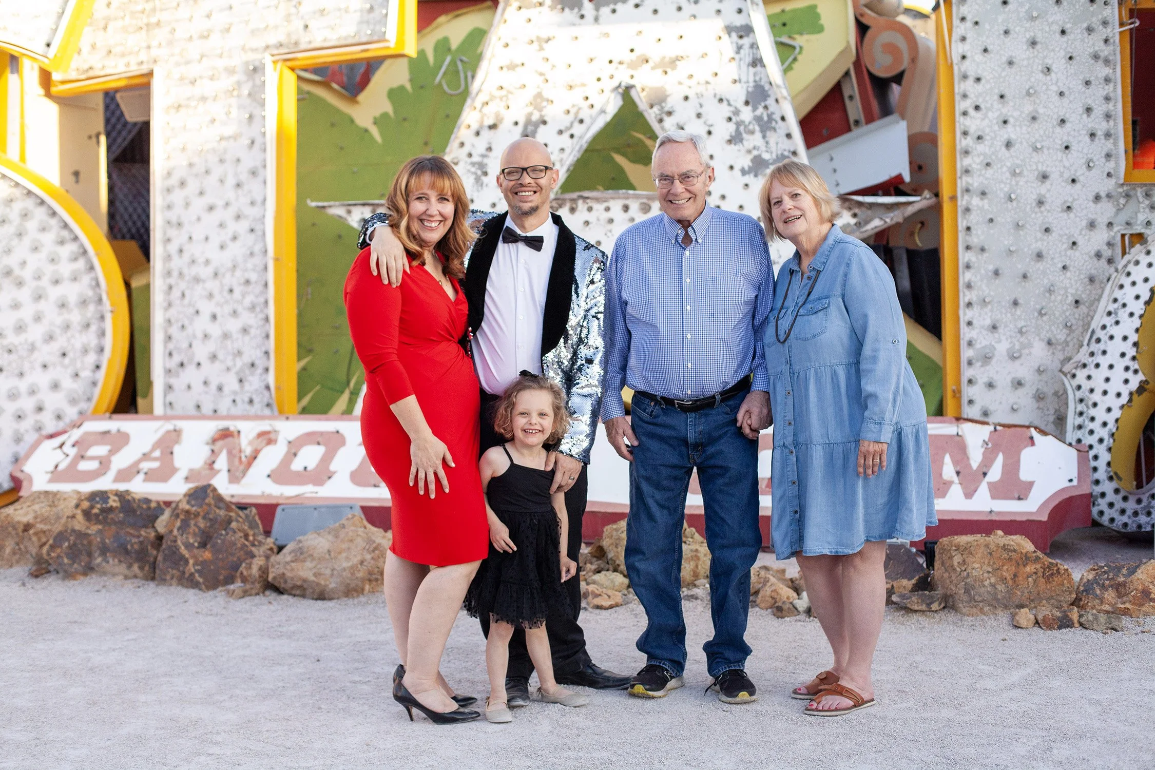 a family of 5, parents, toddler, grandma and grandpa, standing together with large neon signs in the background