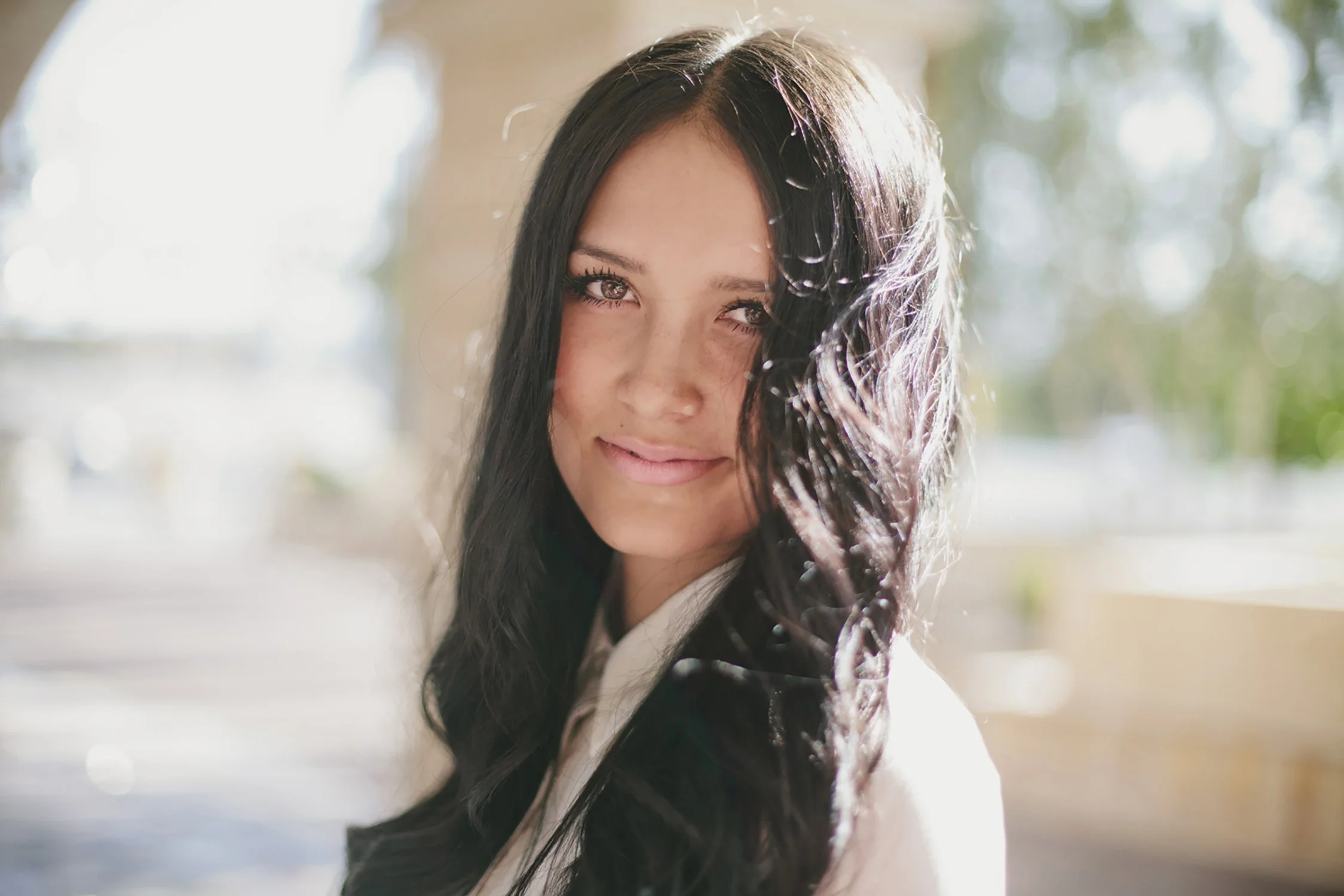 Close up of high school senior, Ashley. She is smiling with her mouth closed looking at the camera with her body facing to the side. The sun is shining off camera on the back of her long hair.