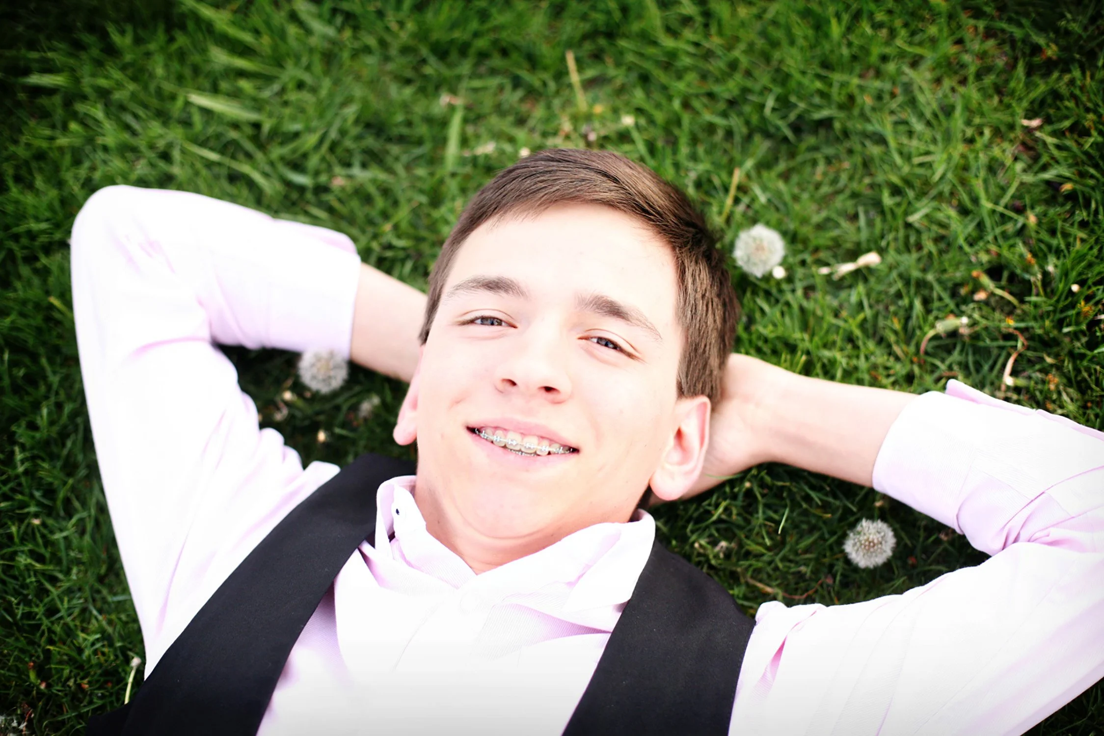 laying on the green grass with his hands behind his head, a high school senior is smiling with braces at the camera.