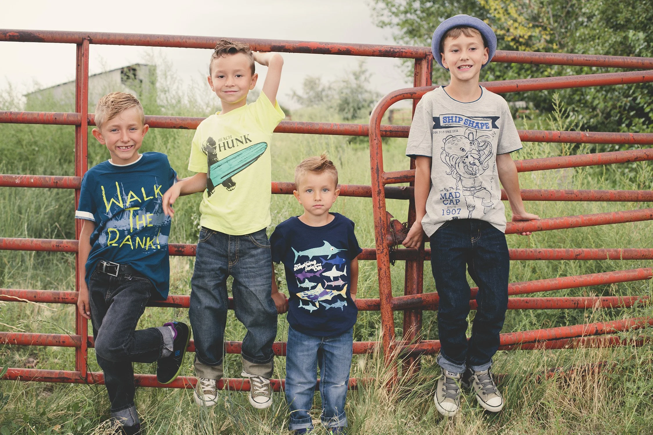 four young brothers, age ranging from 5 to 10 years old, standing on and against a red iron fence with grass and trees in the background