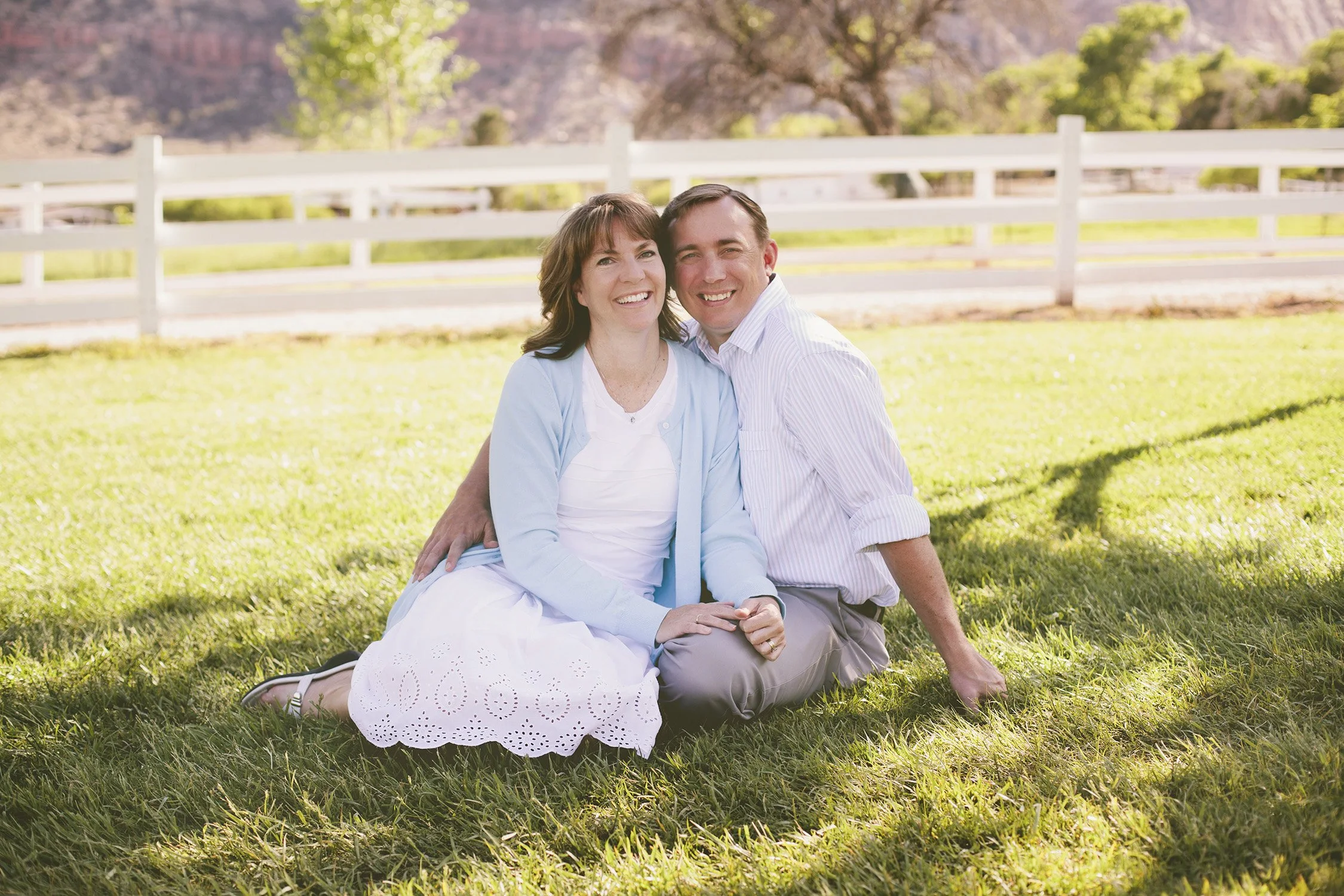 A husband and wife are sitting on the grass close together in the shade of a tree with the mountains, white fence and tress in the background