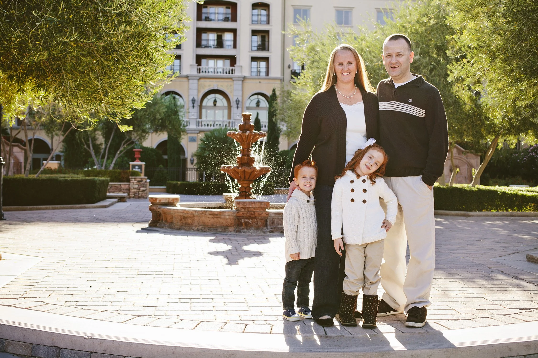 A family of four are standing on a brick pack with a water fountain splashing in the background, included are trees and a large hotel building in the further background. The sun is behind them as they smile looking at the camera.