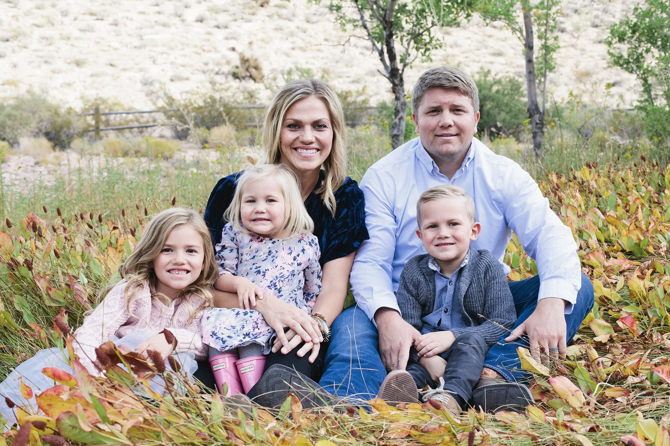 a family of five is sitting on grass and foliage with a mountain, rocks, trees and grass in the background. They are all smiling at the camera. there is a mom and dad, two girls and a boy, age ranging from 3 to 8 years old. Two kids are sitting on mo