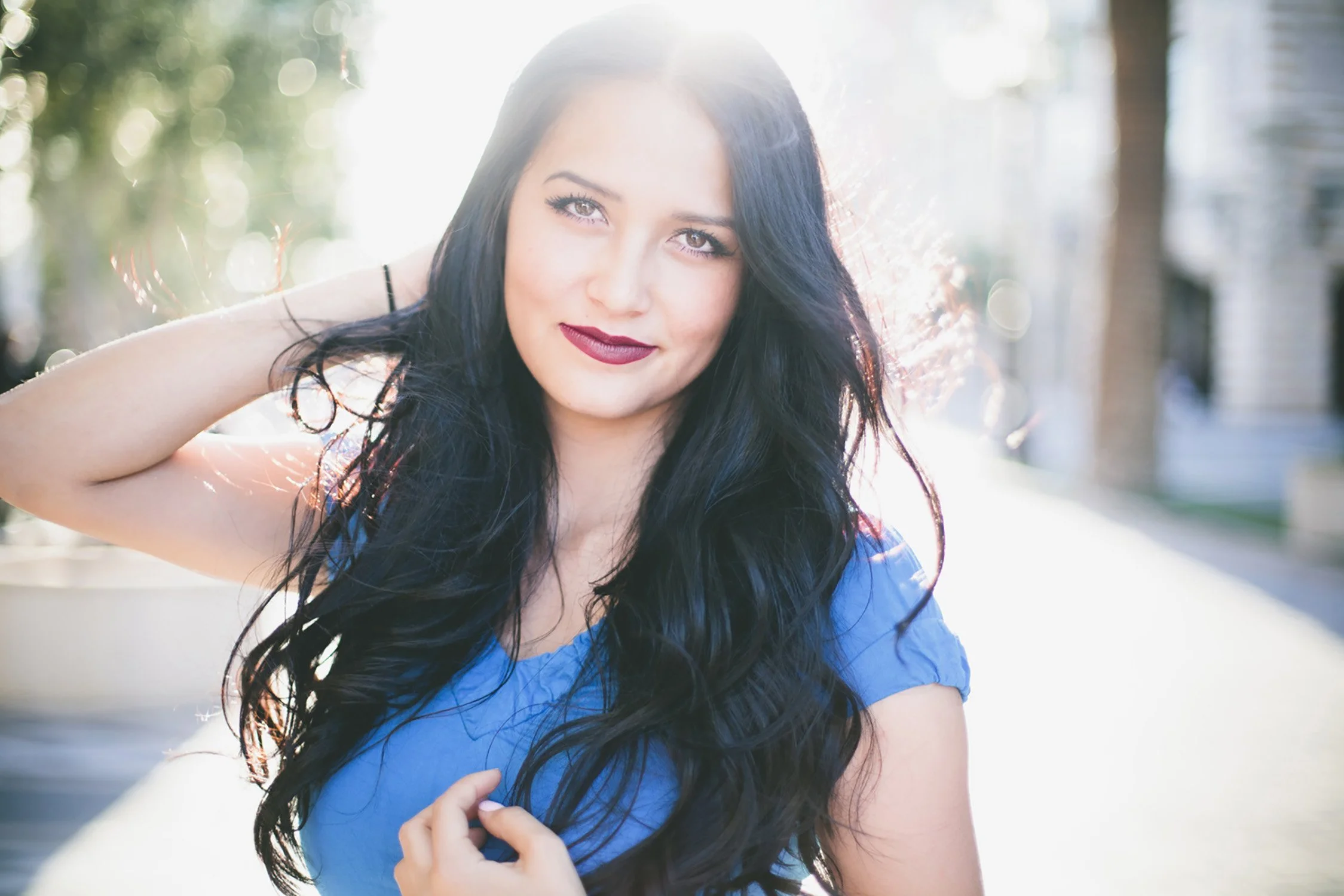 Close up of high school senior, Ashley. The sun is shining behind her, there is a breeze with her long dark hair in the wind, and one hand up towards the back of her head holding her hair back some.