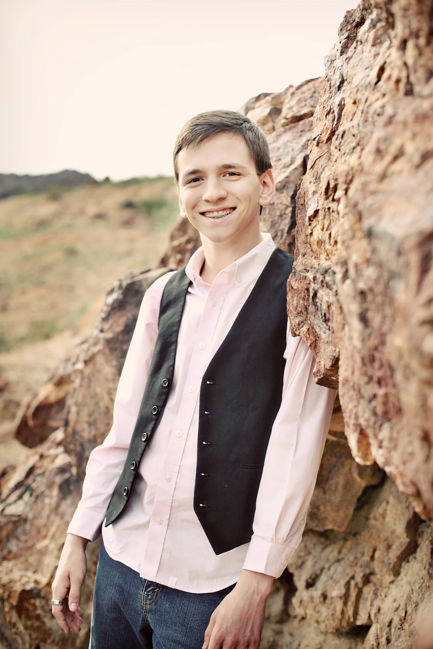 A high school senior is leaning up against some red rocks. He is smiling at the camera