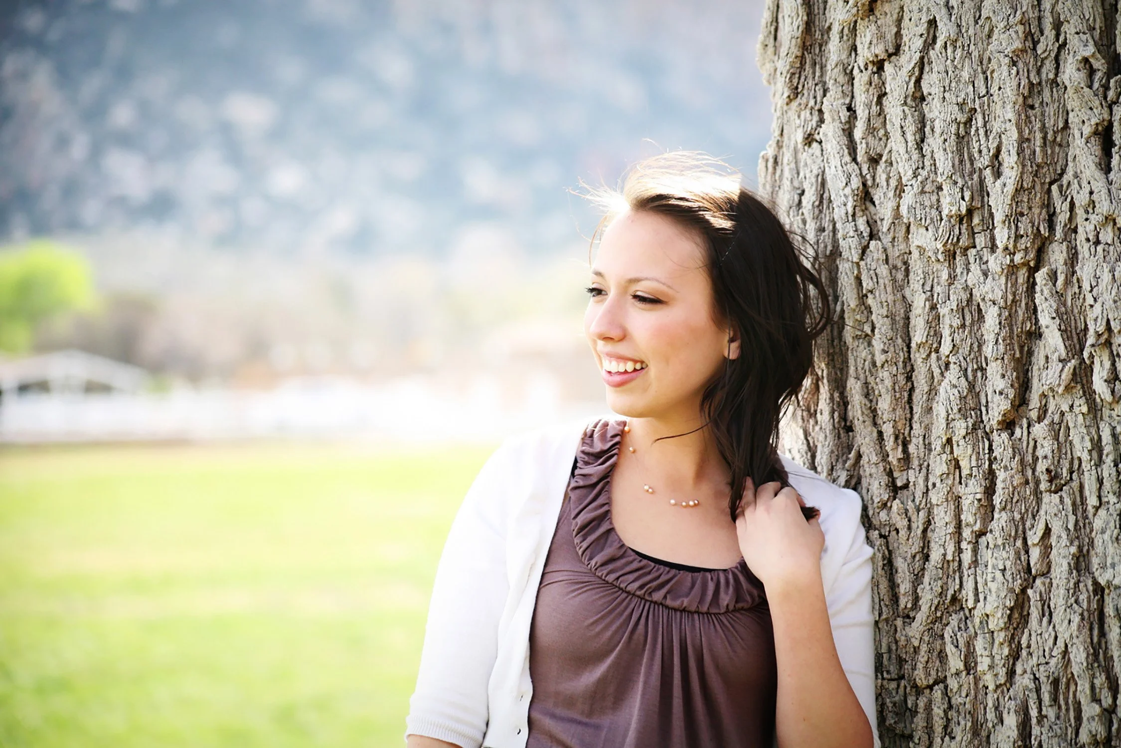 leaning up against a big tree, a high school senior is smiling with teeth and looking away from the camera while holding her hair from blowing in the wind. you can see a green field of grass and mountains in the background.